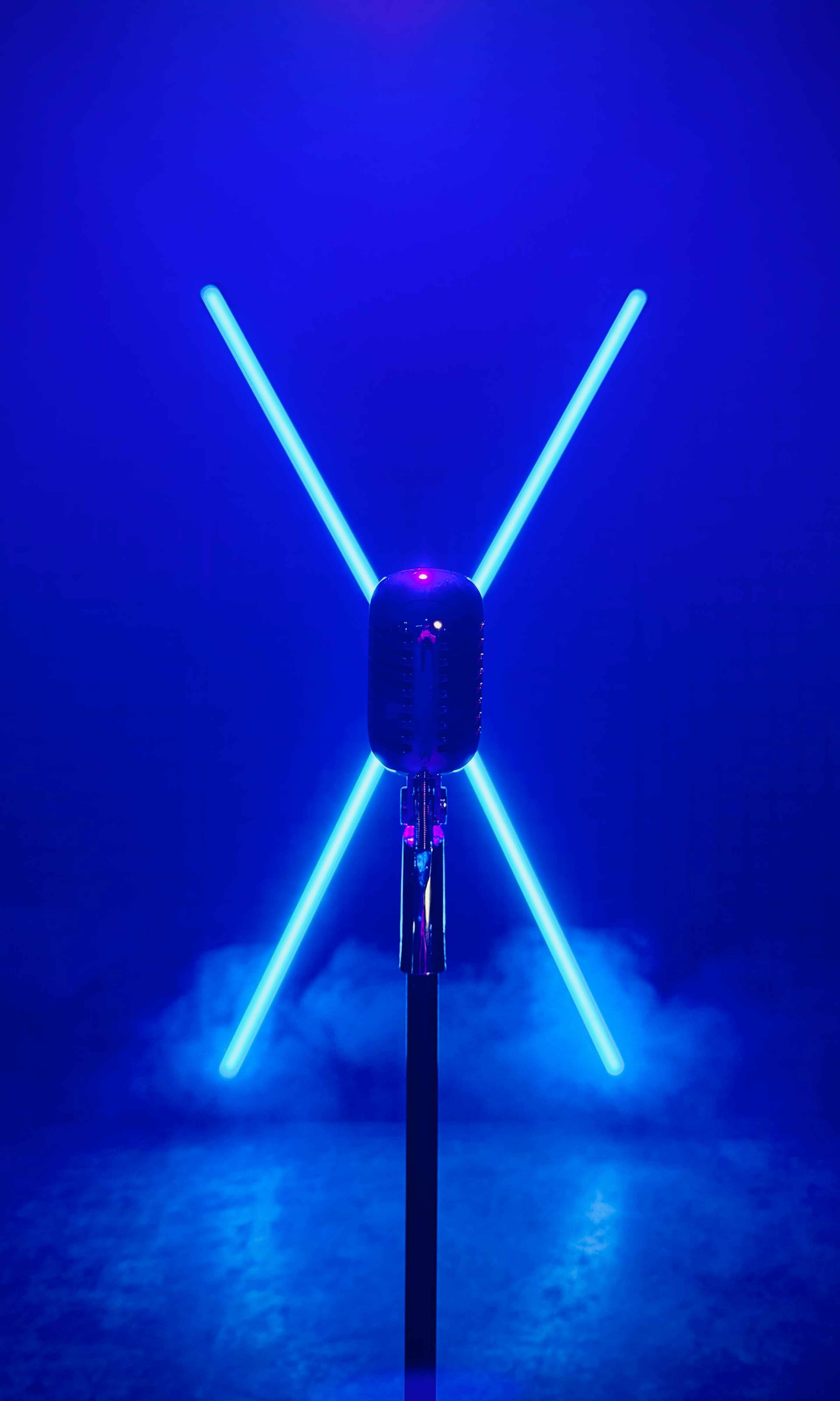 A vintage microphone stands in the center with two bright blue light bars forming an X behind it, surrounded by a light mist.