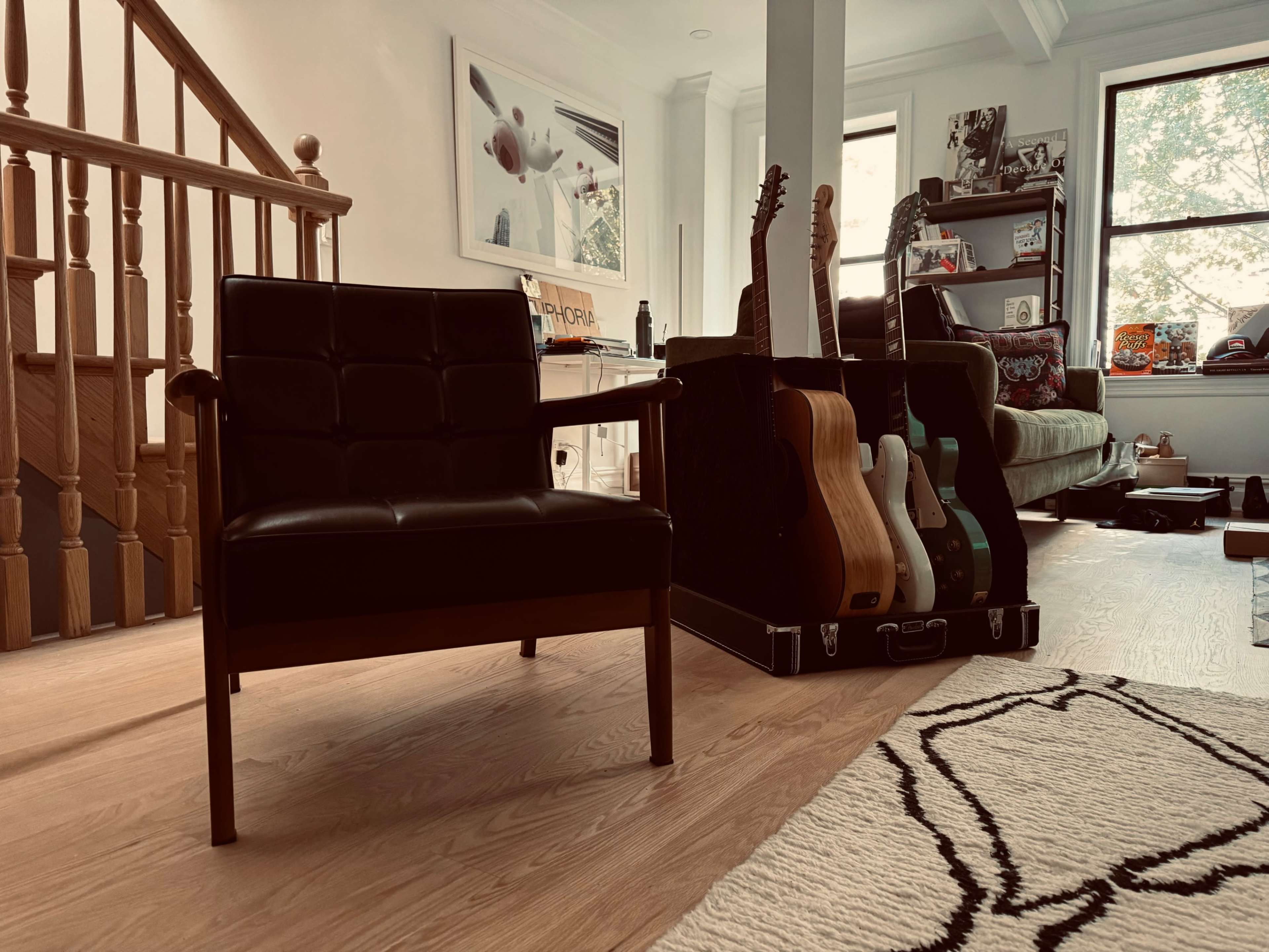 A black armchair is positioned in a living room beside a collection of guitars stored in a case against a wall with windows.