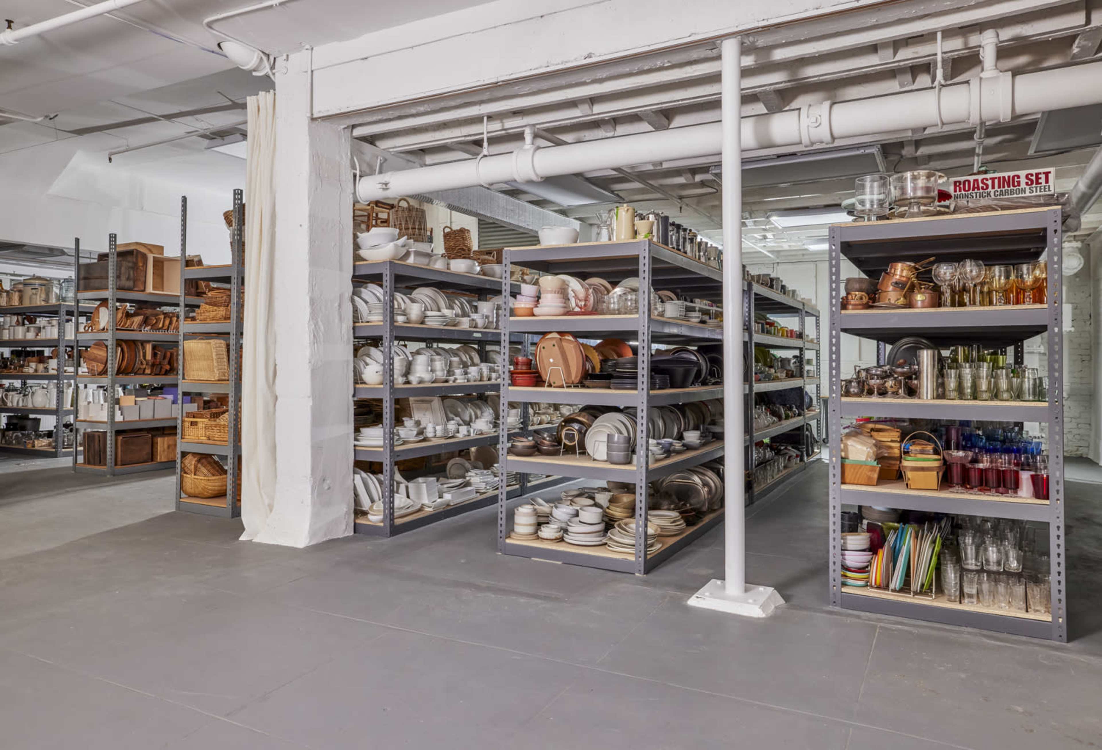 A spacious warehouse filled with metal shelving units displaying various kitchenware, including dishes, serving sets, and storage containers.