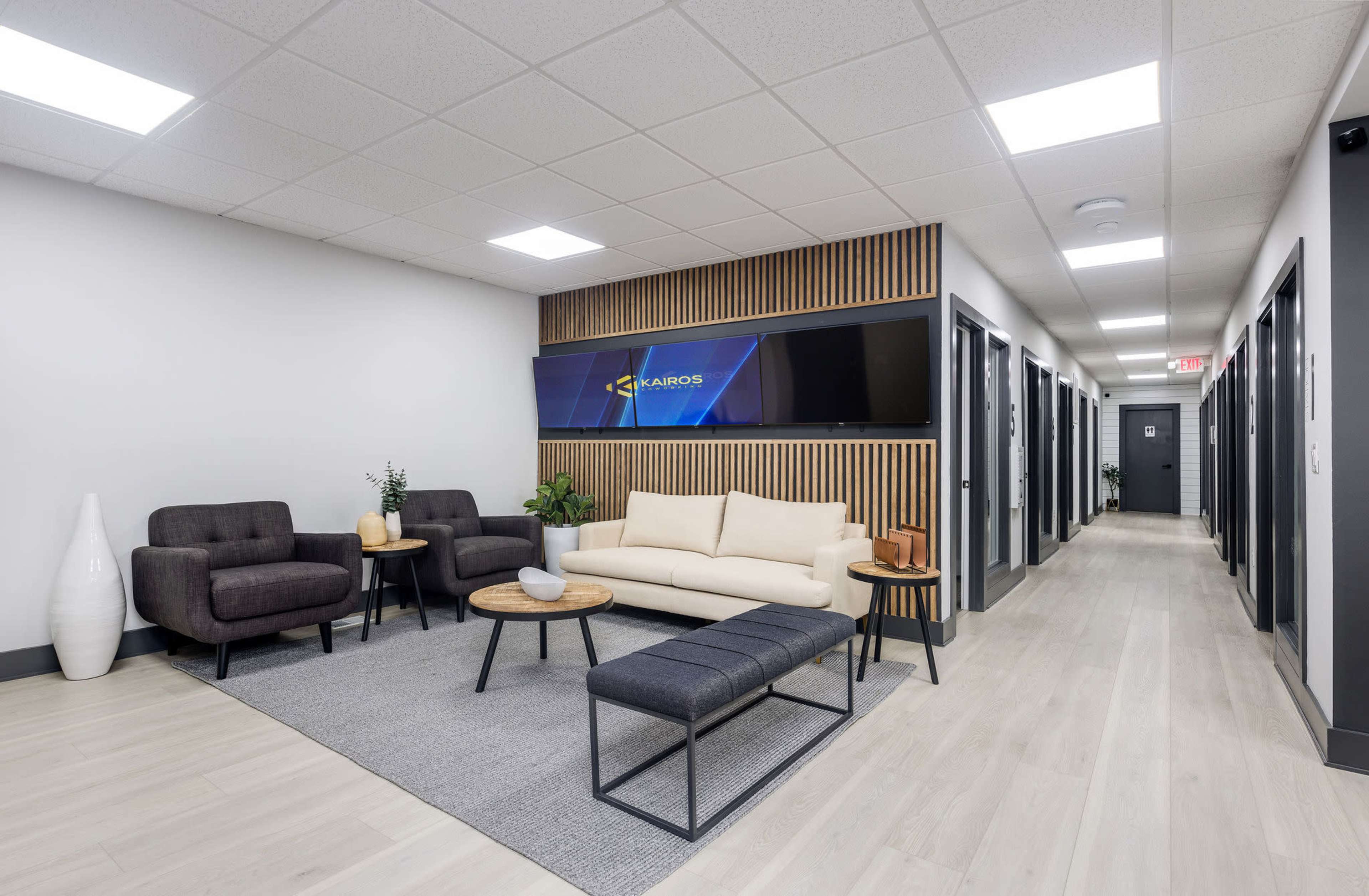 A modern office waiting area with grey sofas, a coffee table, and a large screen mounted on a wooden panel against the wall, leading to a hallway with multiple doors.