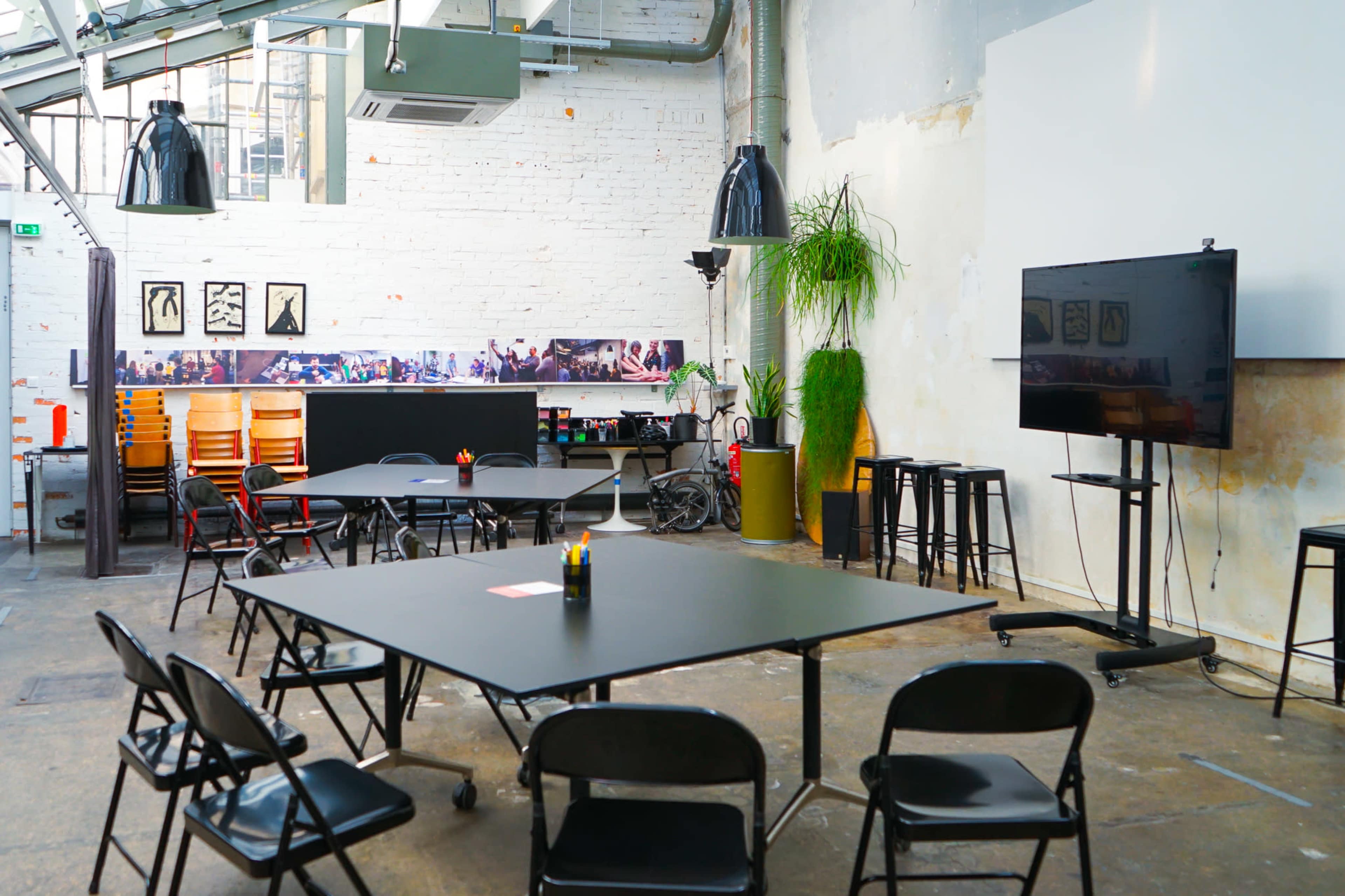 A spacious, industrial-style room features black tables and chairs arranged around a central area, with a large television and a green plant in the background.