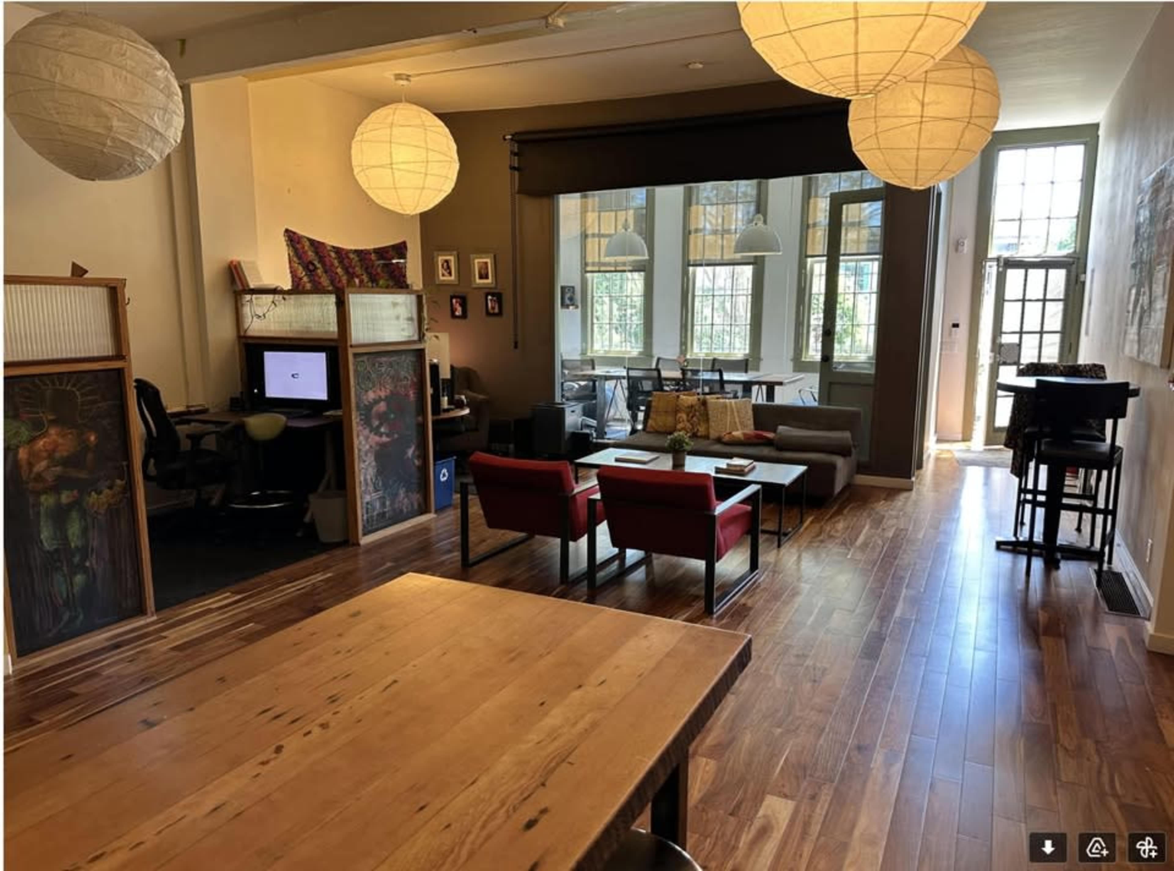 The image shows a spacious room with wooden flooring, a dining table, a seating area with two red chairs, and a workspace separated by a partition.