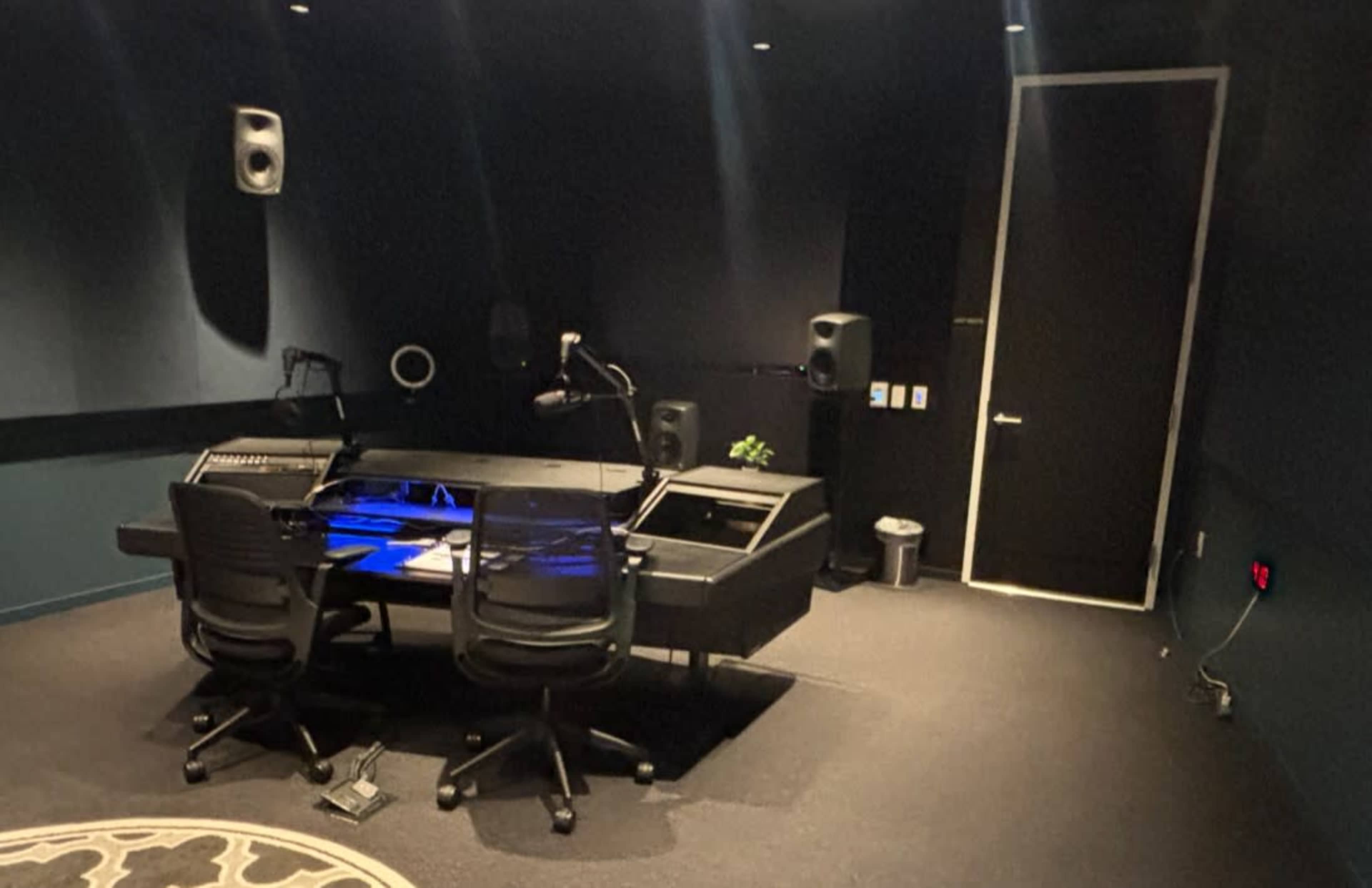 The image shows a recording studio with a black control desk, two office chairs, and surrounding sound equipment in a dimly lit room.