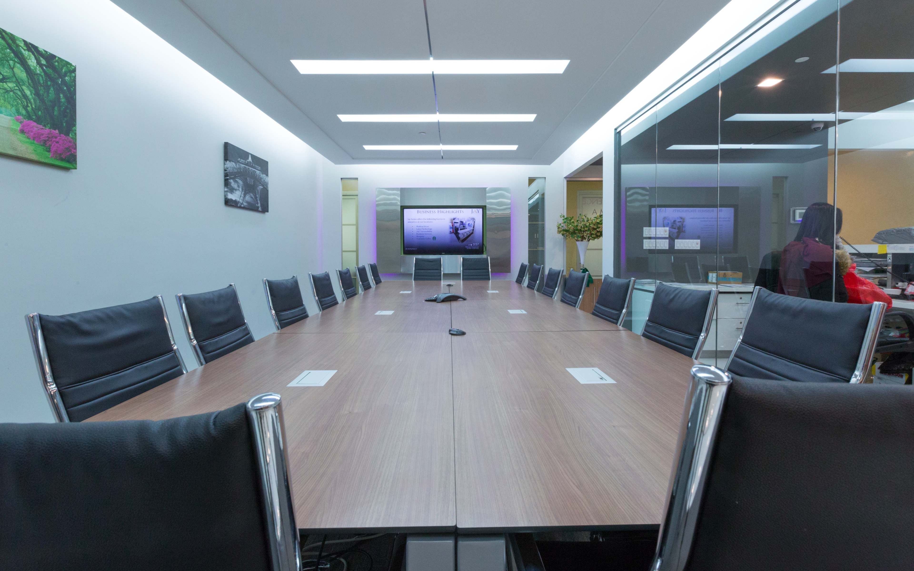 A long, wooden conference table with black leather chairs is positioned in a bright meeting room featuring two screens on the walls.