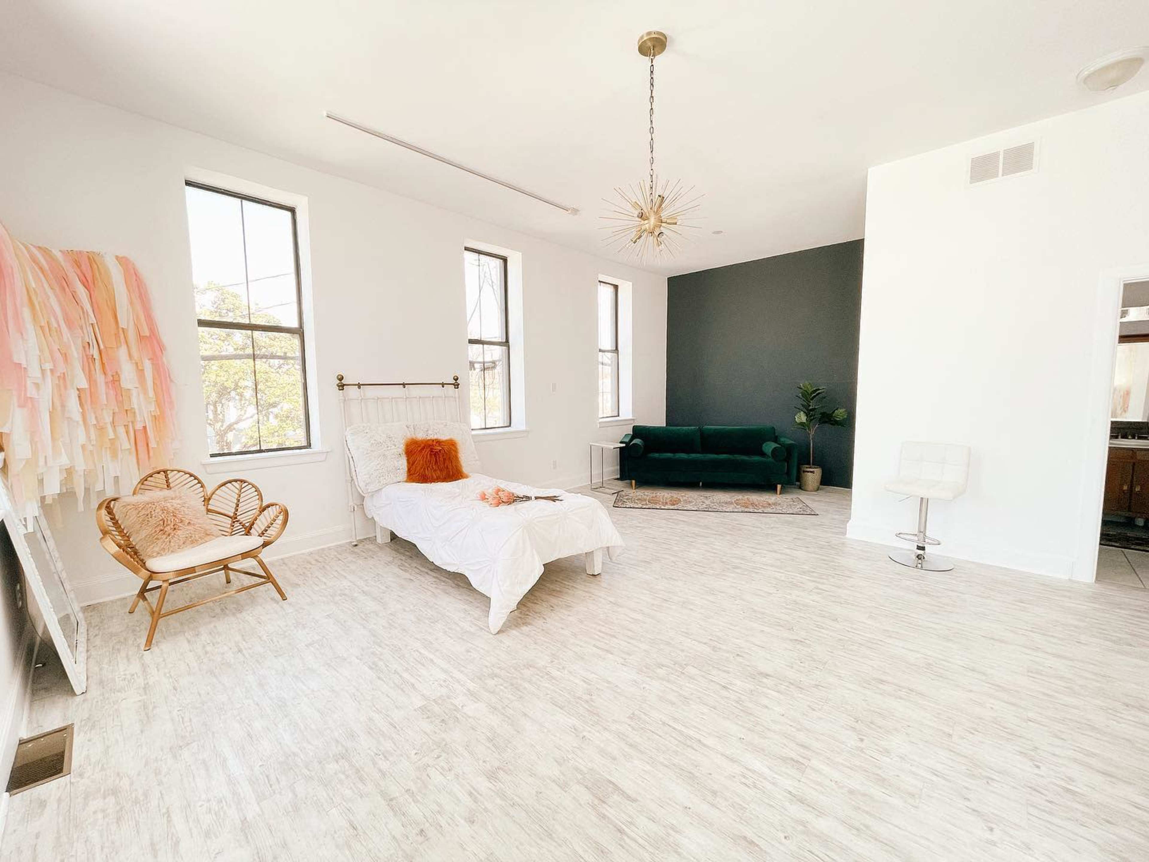 The scene shows a bright, minimalist room with a white bed, a rattan chair, a green sofa, and decorative wall elements.