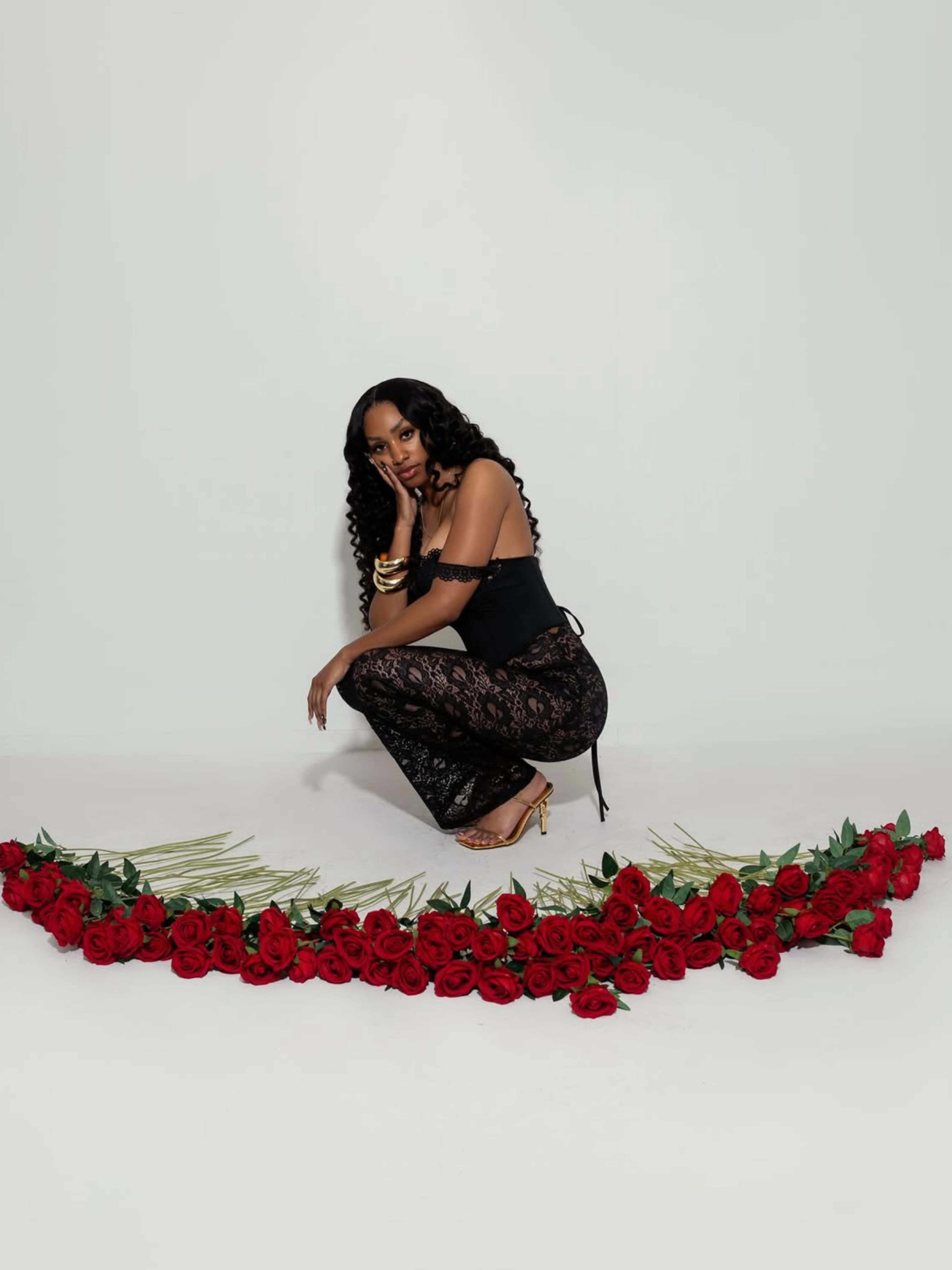 A woman in a black outfit squats beside a semicircle of red roses on a light background.
