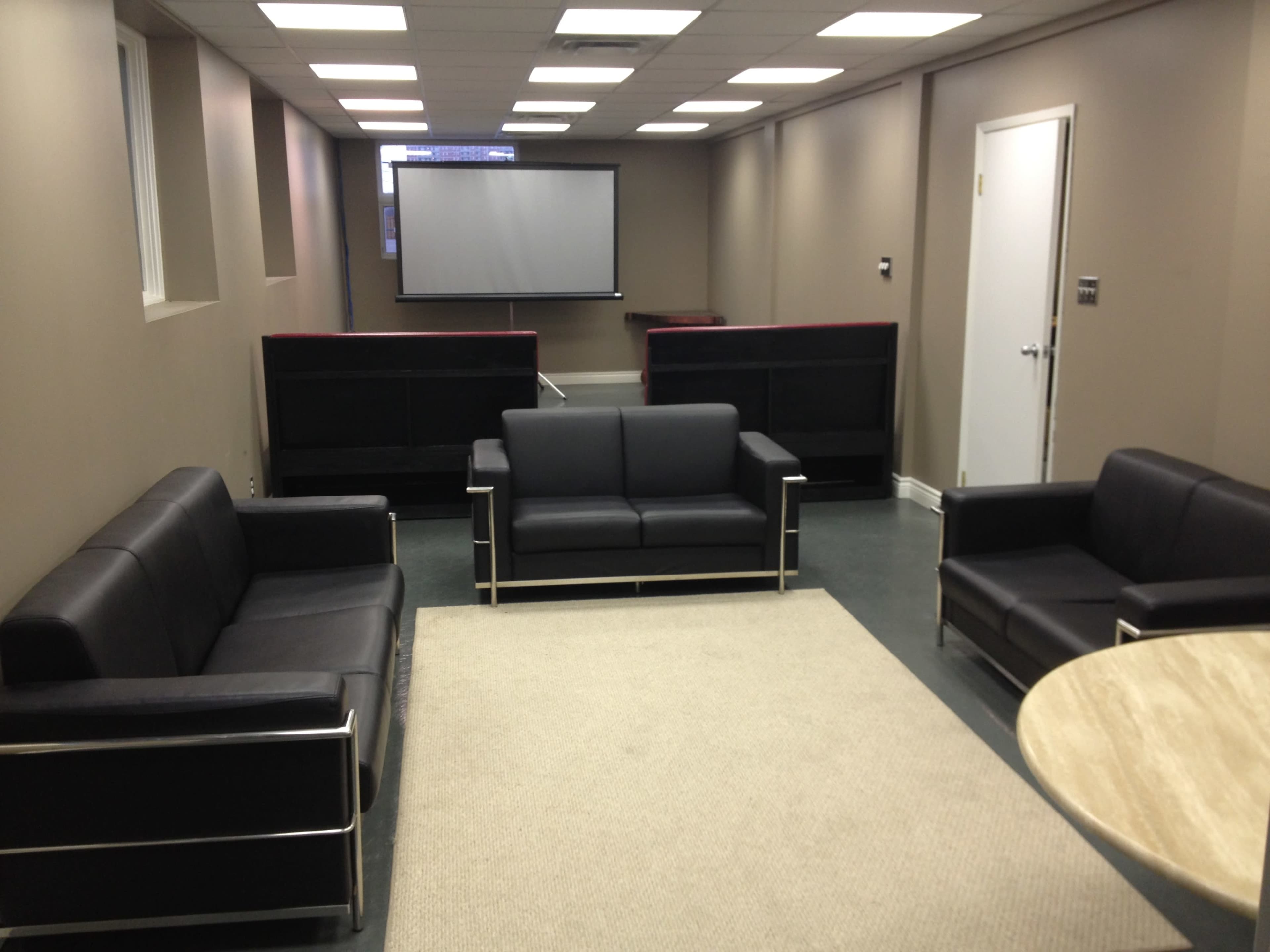 A sparsely furnished room with three black leather couches, a large screen at the front, and a round table on the side.