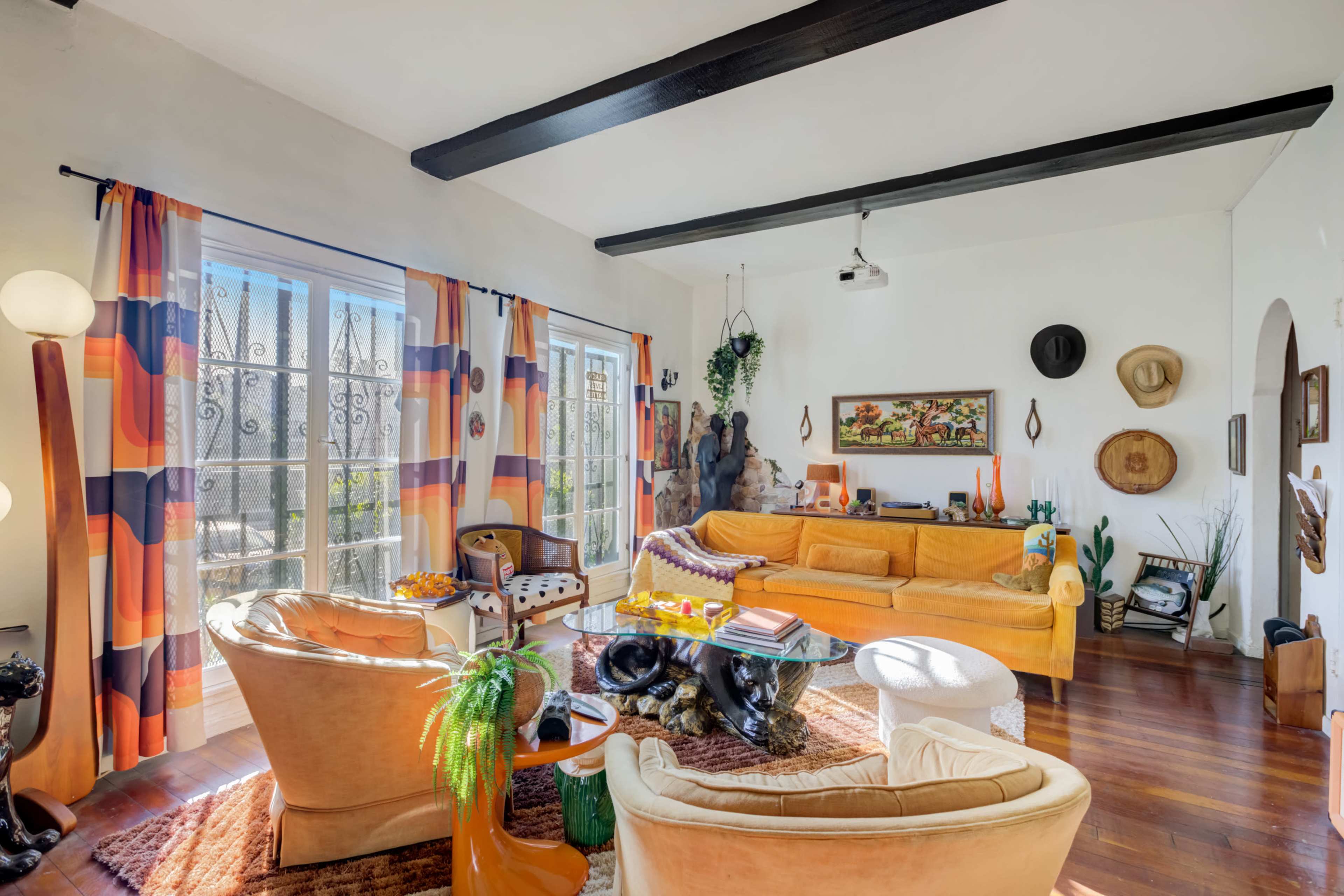 The living room features a yellow sofa, large windows with colorful curtains, and various decorative items, including a sculpture and potted plants.