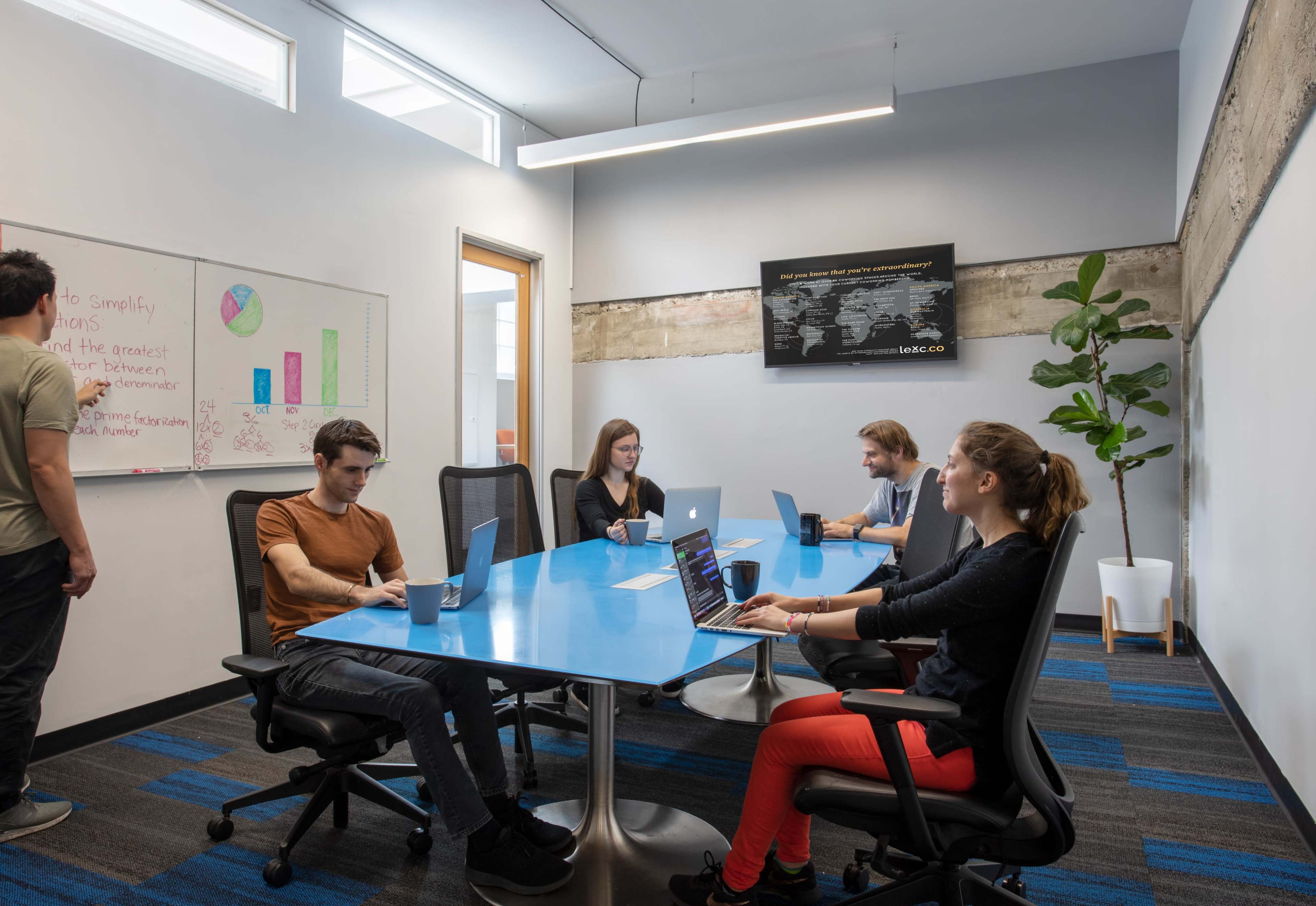 A group of four people work at a blue conference table in a modern office, with laptops and a whiteboard in the background.