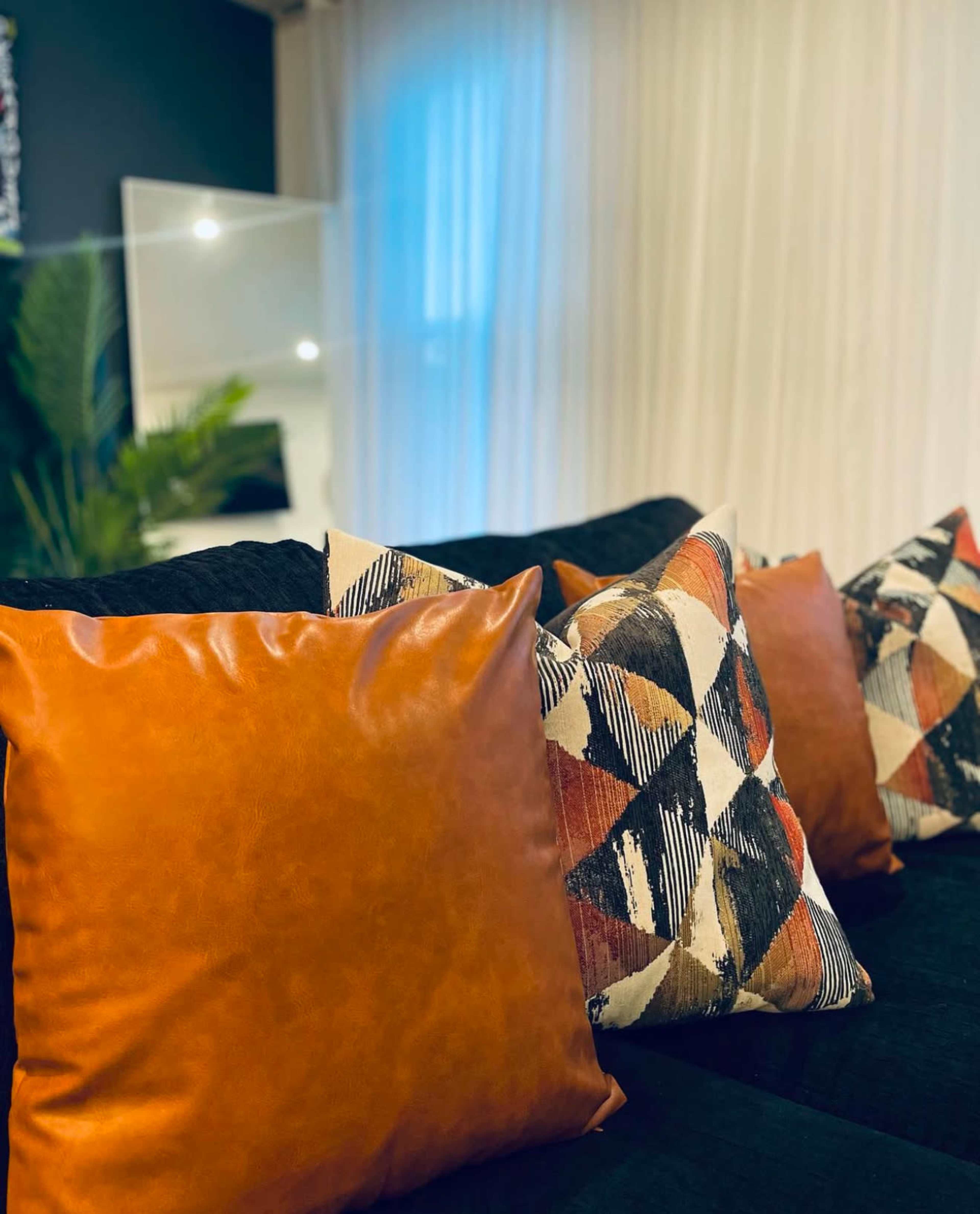 The image shows a close-up of a couch with several decorative pillows, including one leather pillow and others with geometric patterns, set against a backdrop of a light-colored curtain and a mirror.