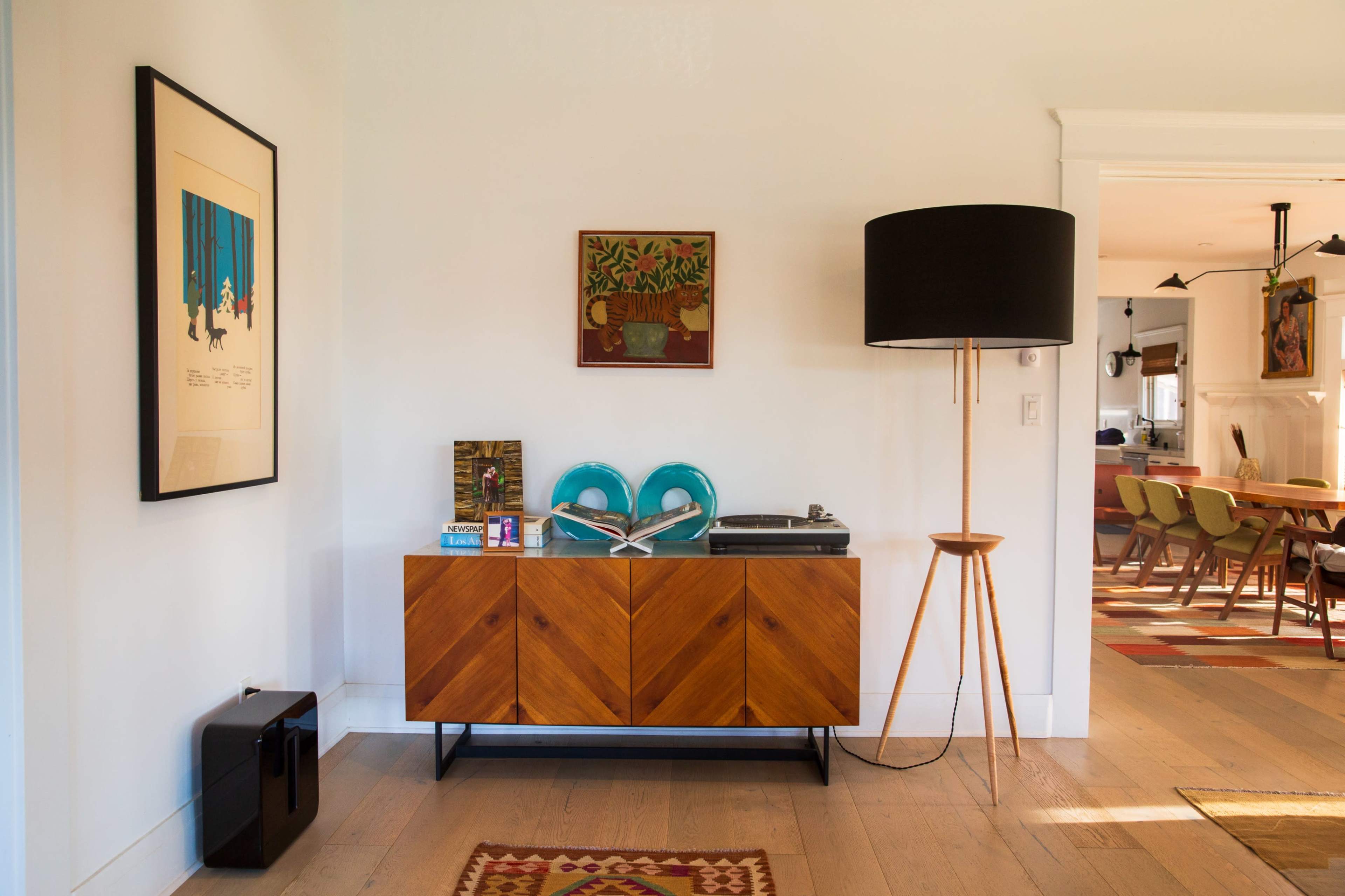 The image shows a modern living space featuring a wooden sideboard with a record player, decorative items, and framed art on the wall, accompanied by a tall floor lamp and an open dining area in the background.