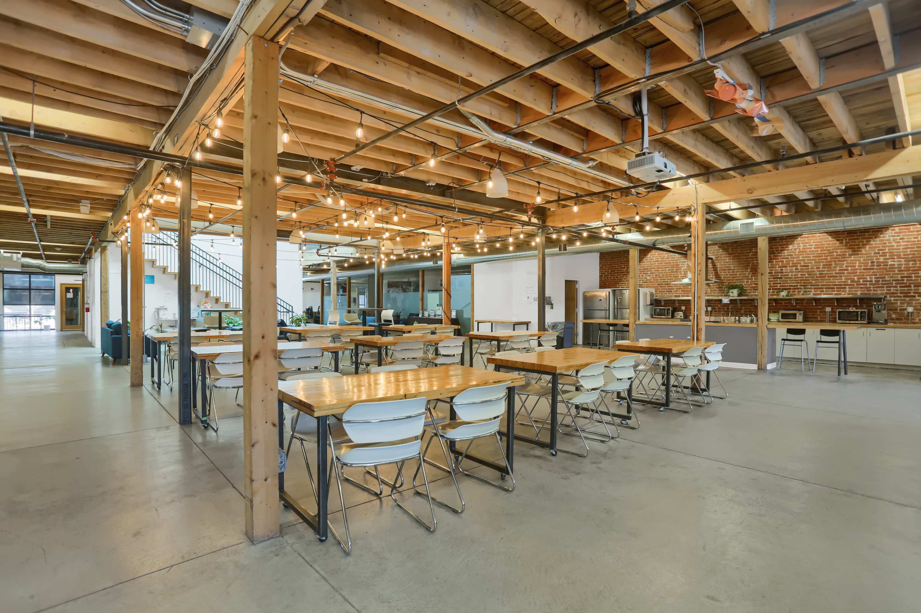 The image shows a spacious, well-lit industrial-style interior with wooden beams and multiple dining tables surrounded by chairs.