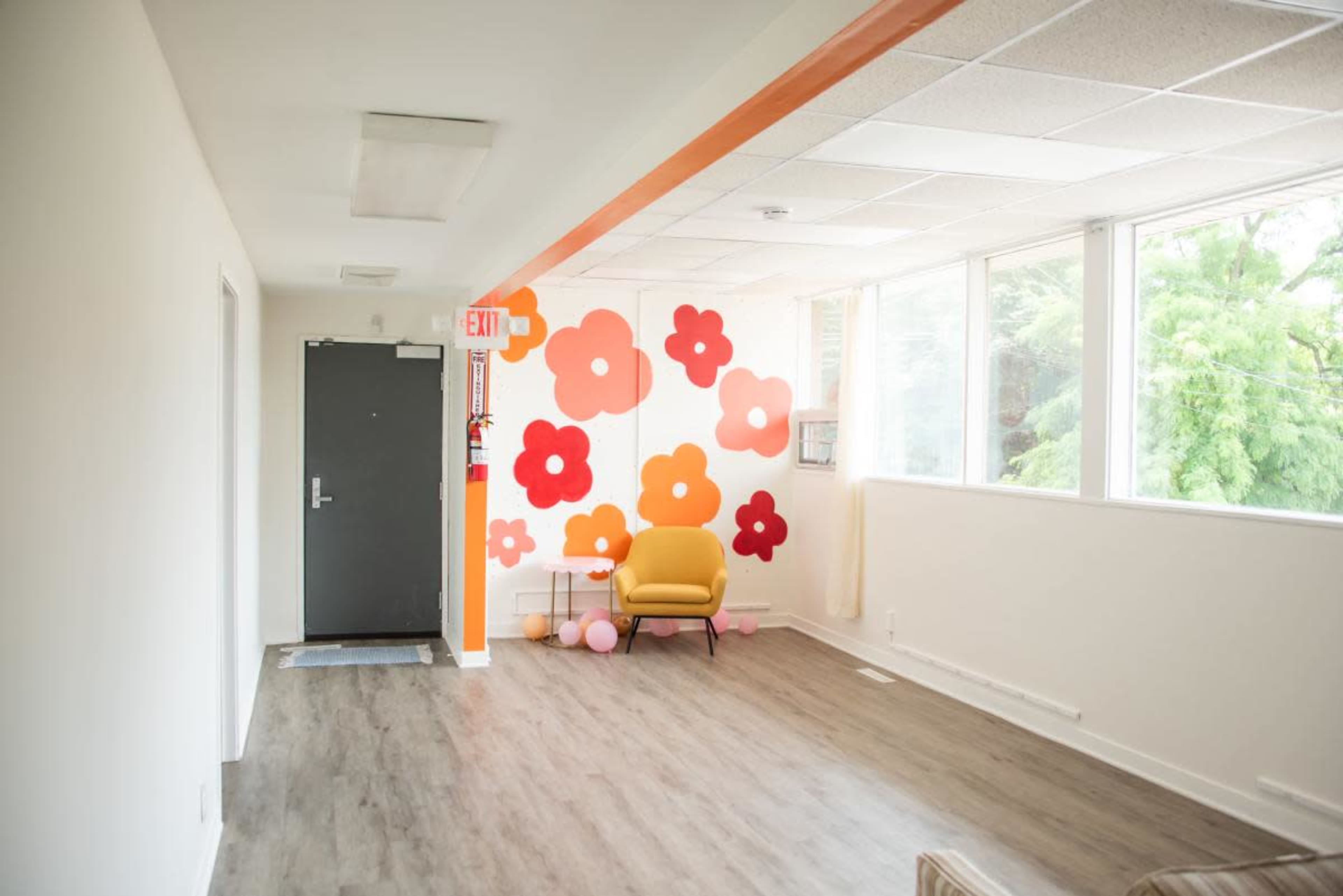 The image shows a brightly lit hallway with a colorful flower mural on one wall, an open door leading to the outside, and a yellow chair beside a small cluster of balloons.