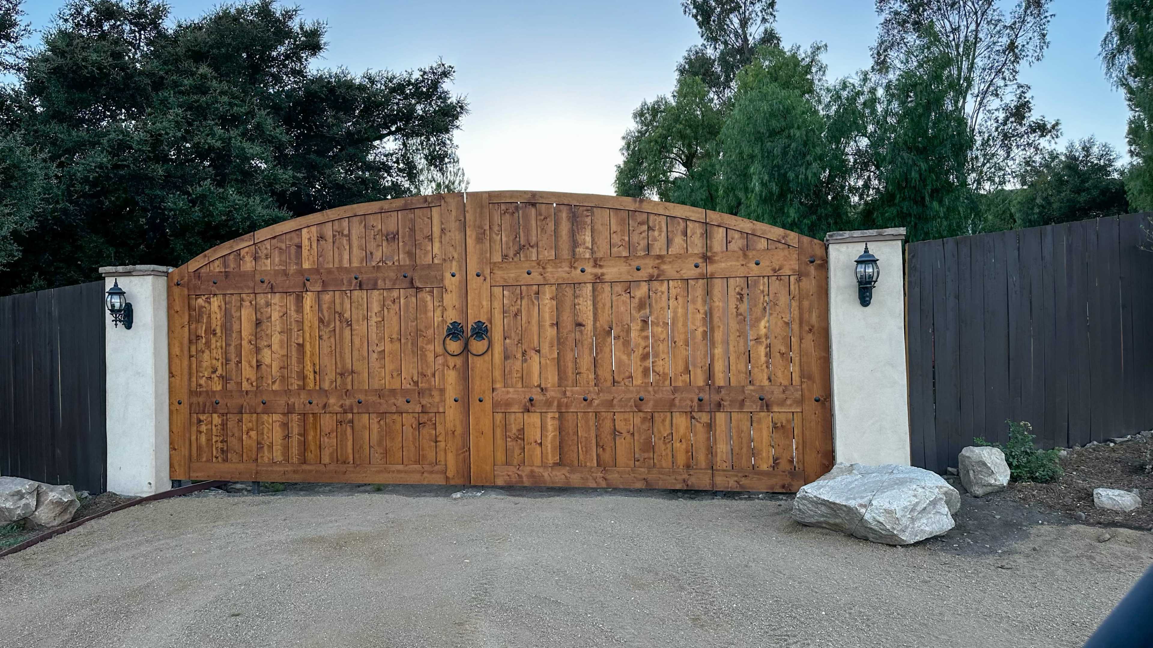 A large wooden double gate with black hardware and lanterns on either side stands at the entrance of a gravel driveway, surrounded by greenery.