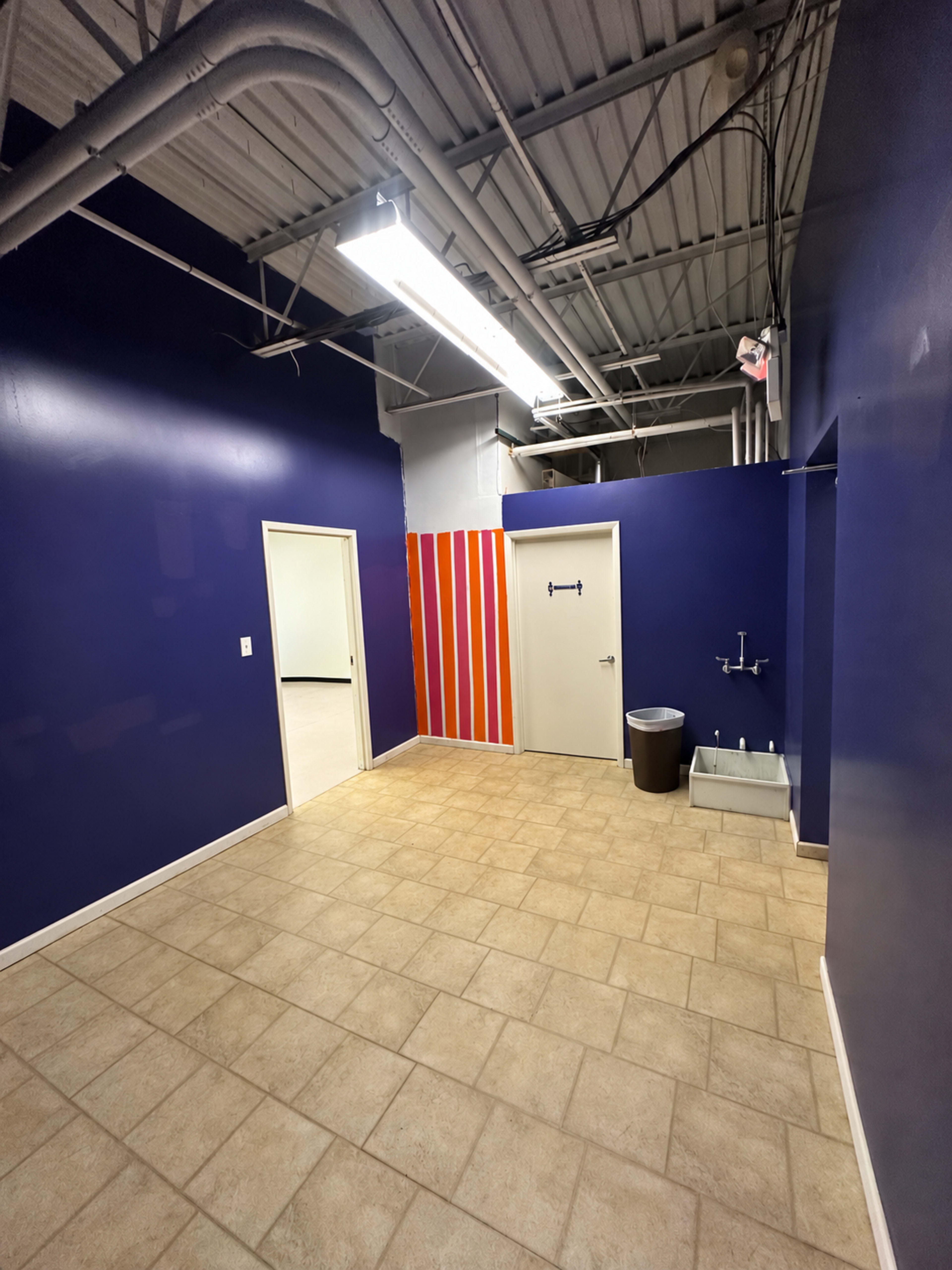The image shows an empty room with purple walls, beige tiled flooring, two doors, and a drinking fountain on one side.