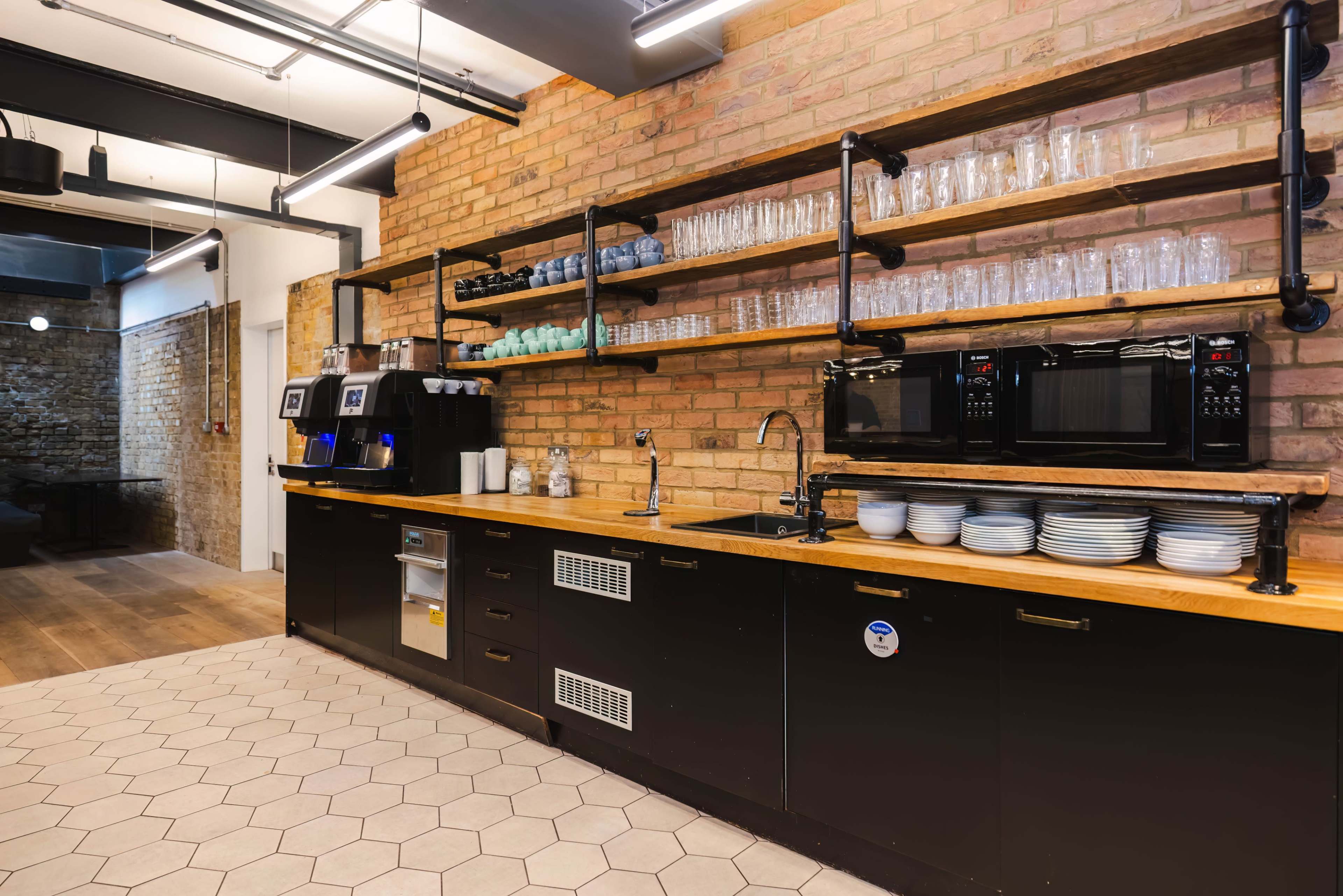 The image shows a modern kitchen with a brick wall, wooden shelves stocked with glassware, and appliances including coffee machines and microwaves.