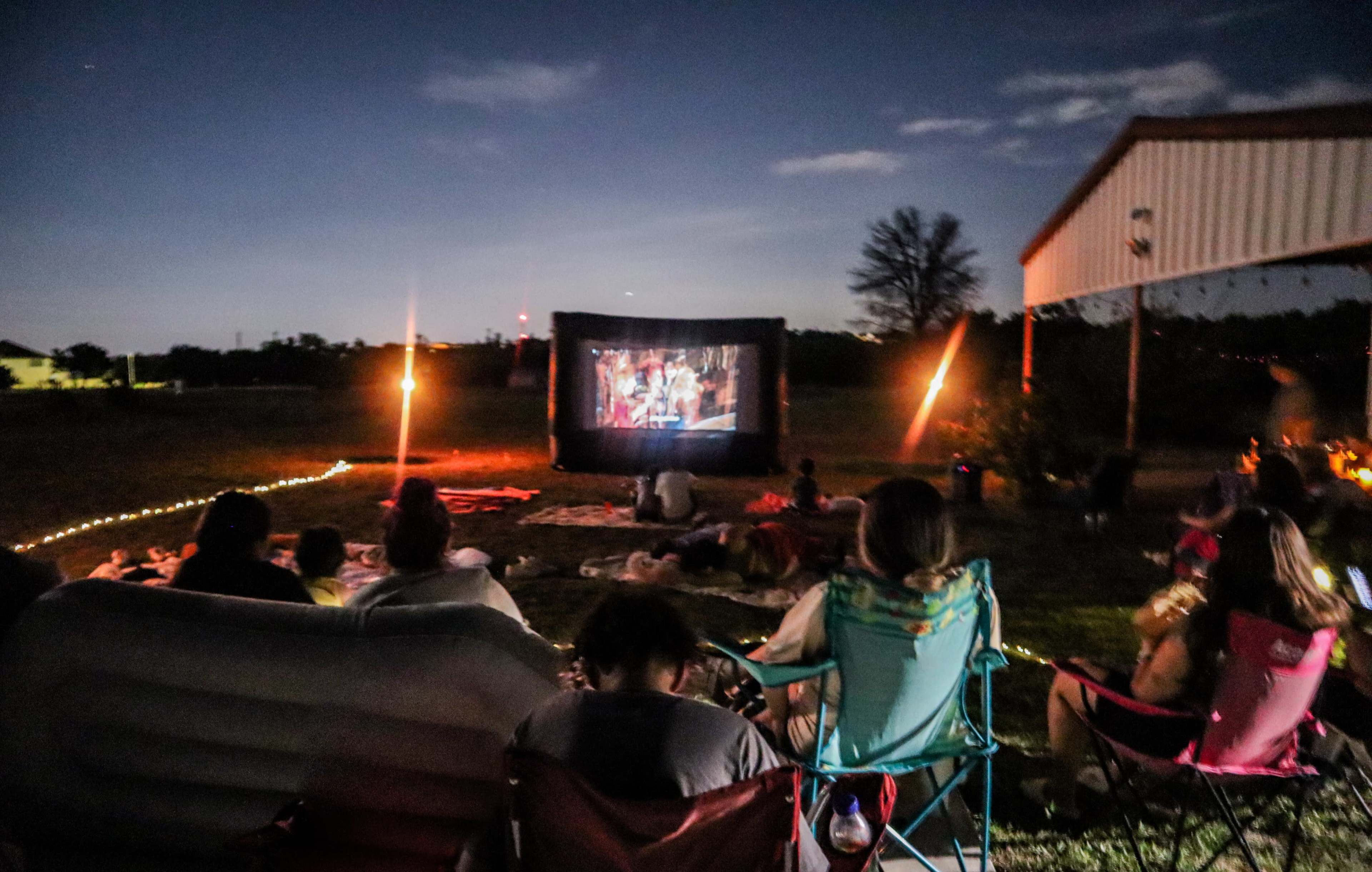 A group of people watch a movie on a large outdoor screen in a field during the evening.