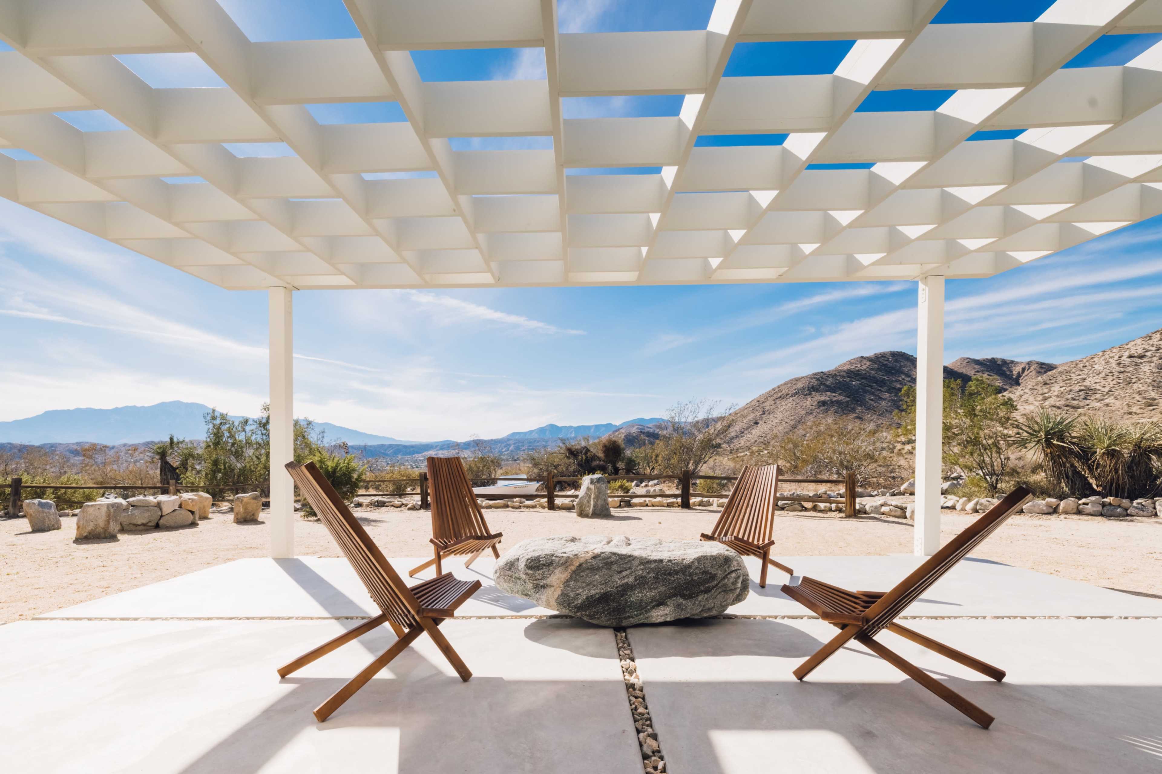 A modern outdoor seating area features four wooden chairs arranged around a large stone in a desert landscape with mountains in the background.