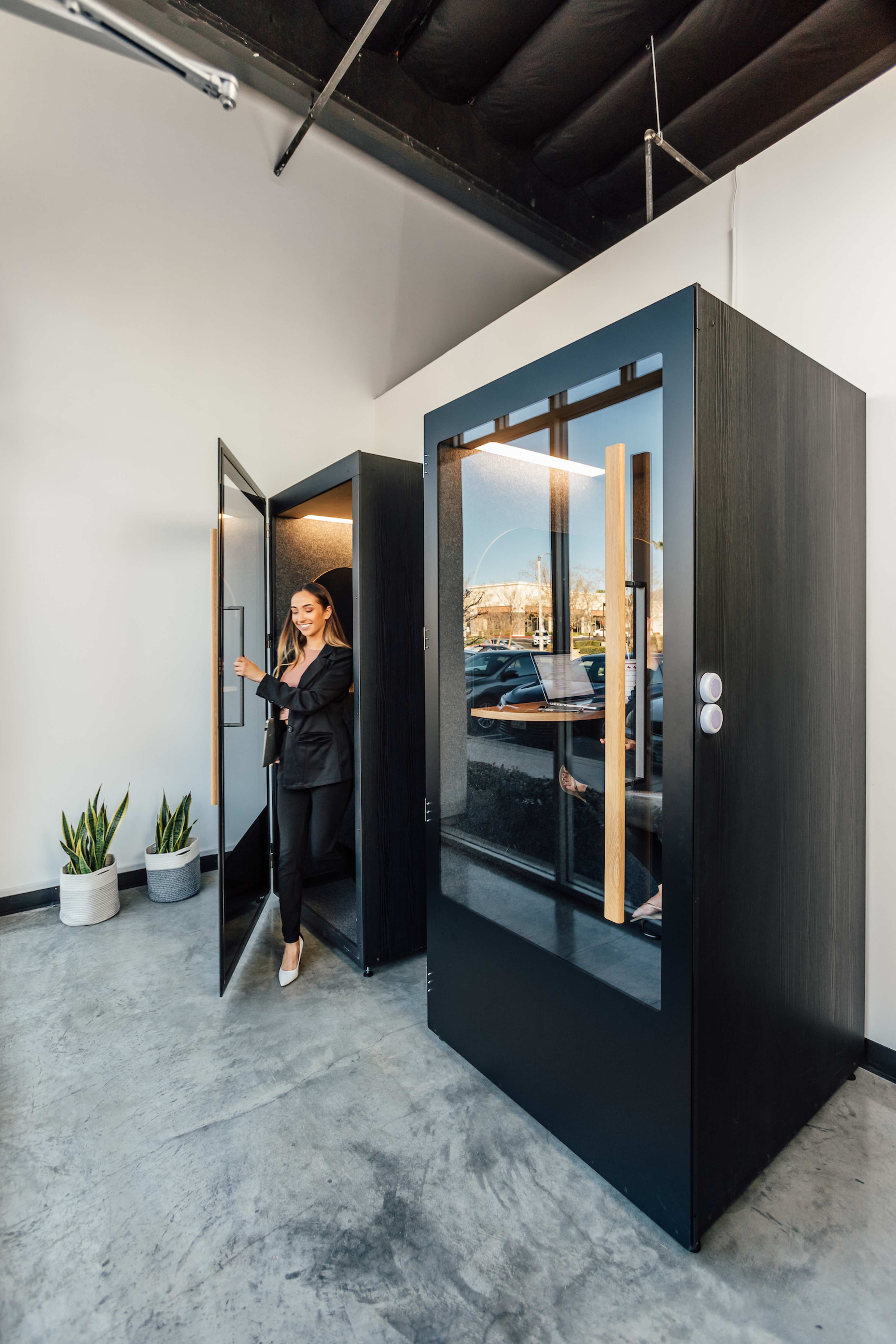 A woman exits a sleek, black phone booth in a modern office space, while another booth stands nearby with its door open.