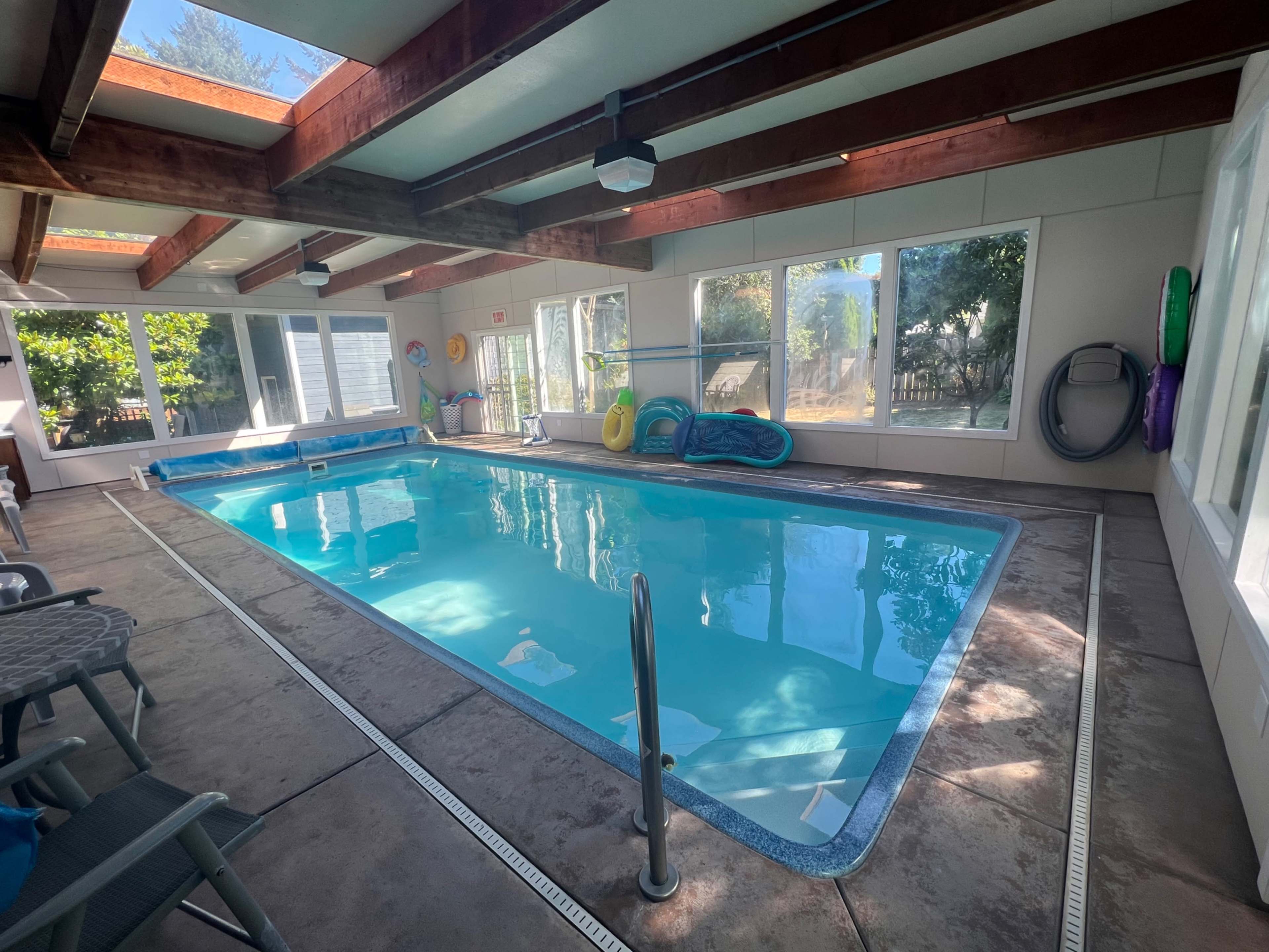 An indoor swimming pool surrounded by large windows and wooden beams, with poolside furniture and colorful floaties nearby.