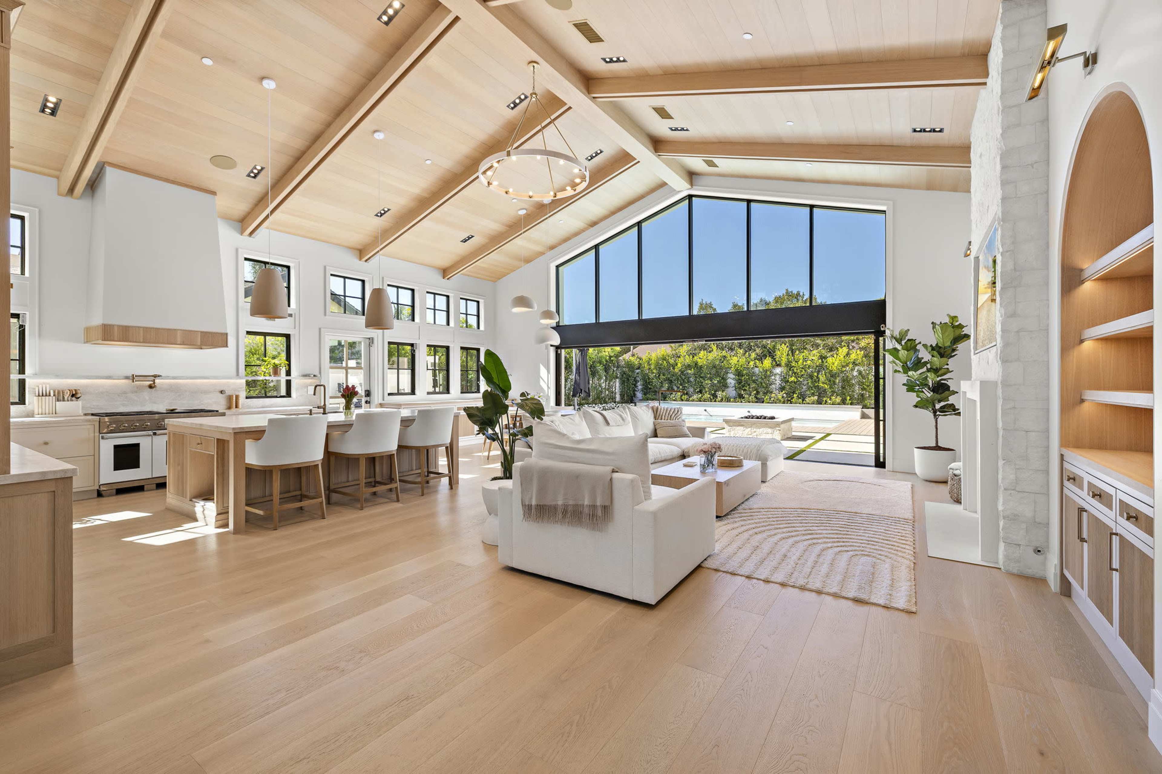 The image shows a spacious, modern living area with high wooden ceilings, large windows, and a light color palette, featuring a cozy seating arrangement and an open kitchen space.