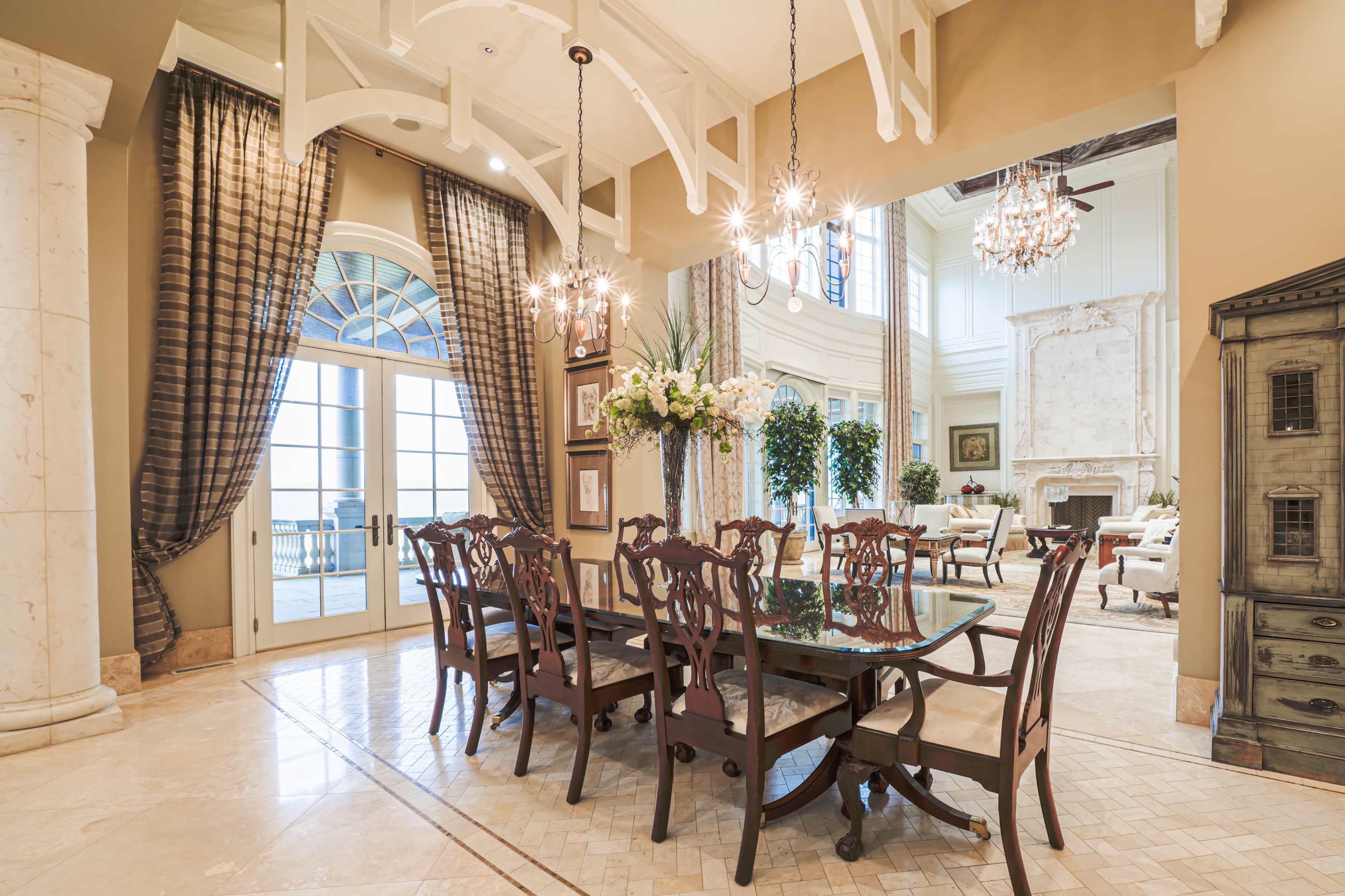 A formal dining room features a large wooden table surrounded by upholstered chairs, with elegant curtains framing tall windows that lead to a view of a living area.