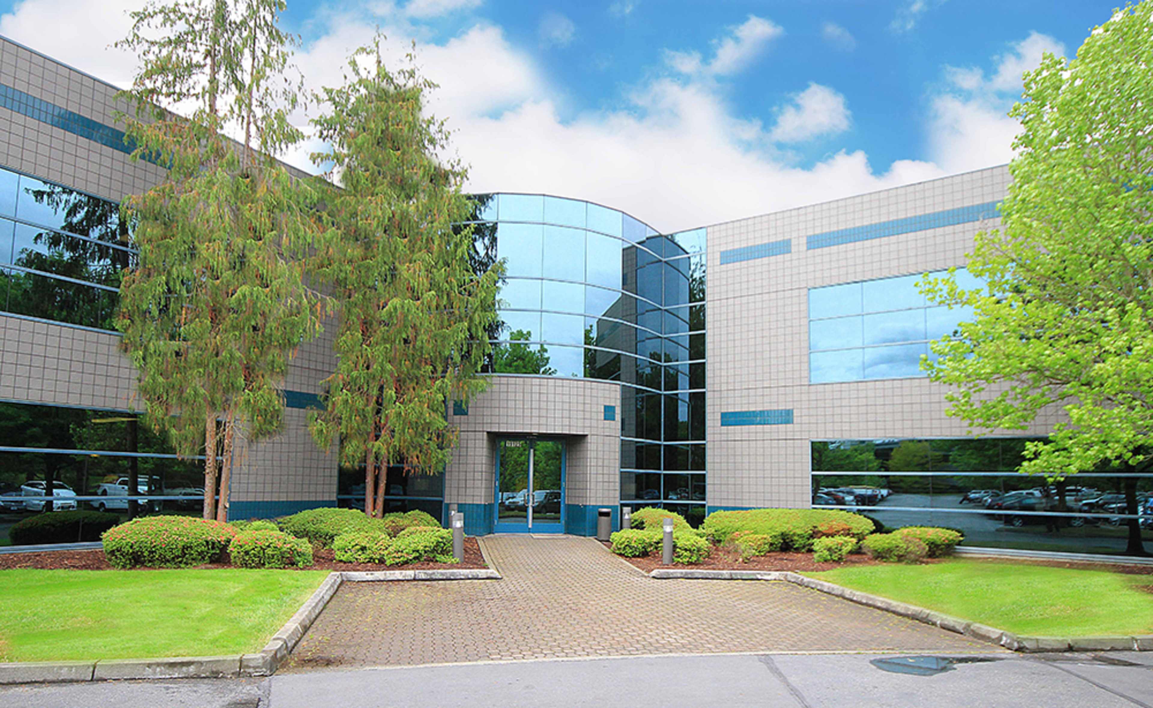 The entrance of a modern office building with large glass windows, landscaped greenery, and a paved walkway.