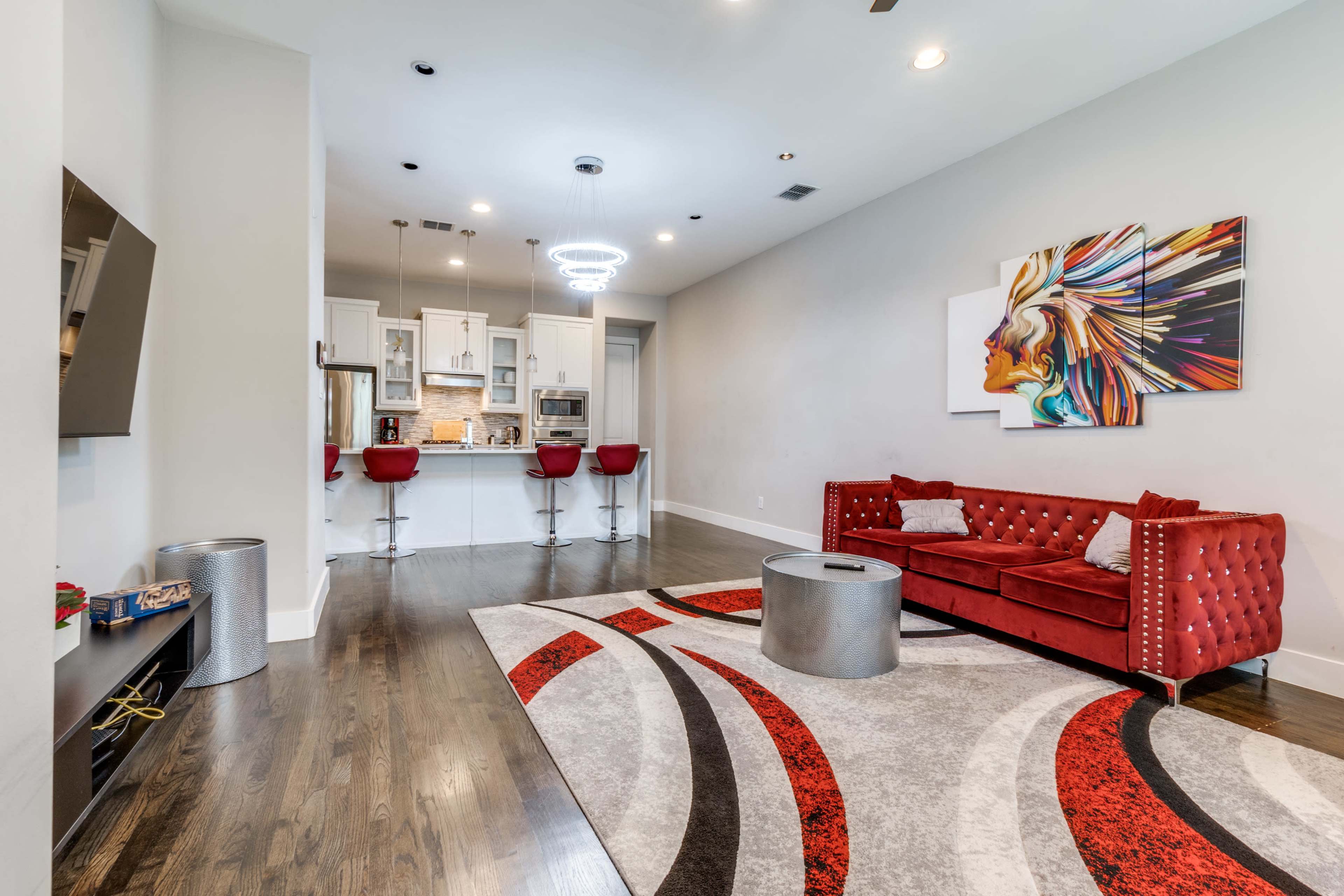 The image shows a modern living space with a red tufted sofa, a circular silver coffee table, and a kitchen area in the background featuring bar stools and contemporary cabinetry.