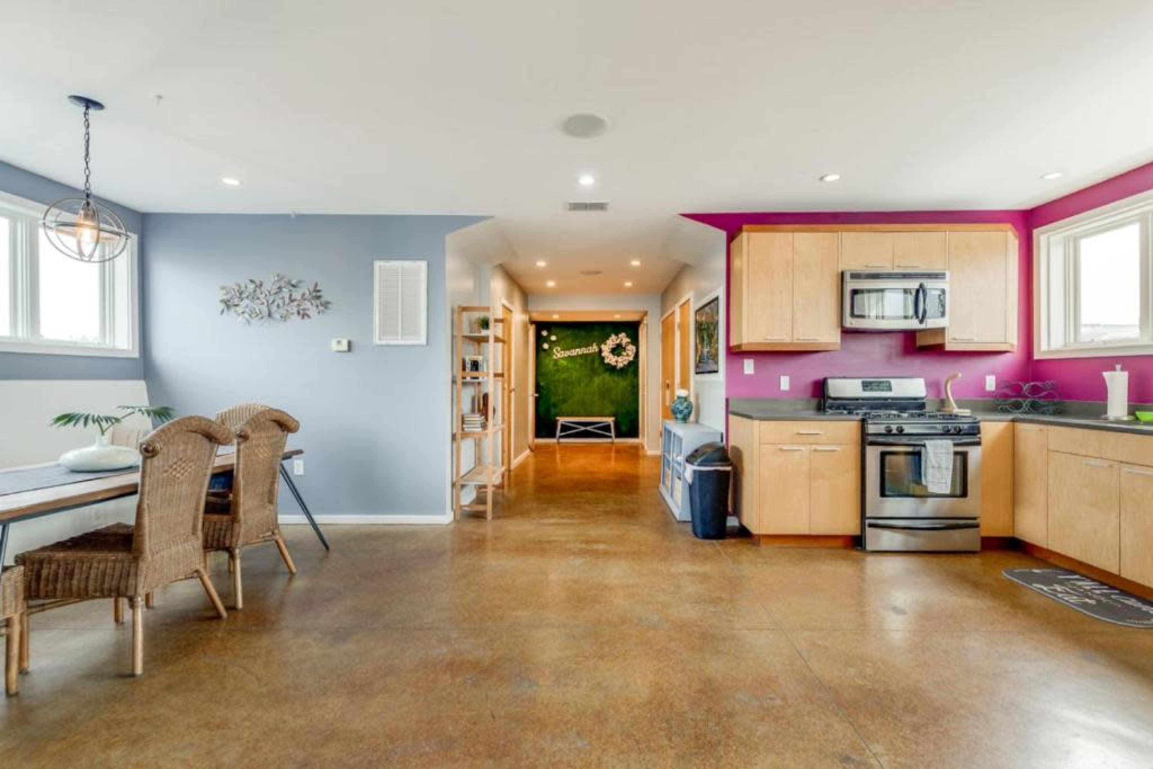 The image shows an open-concept living space featuring a kitchen with stainless steel appliances, a dining area with wicker chairs, and bright accent walls.
