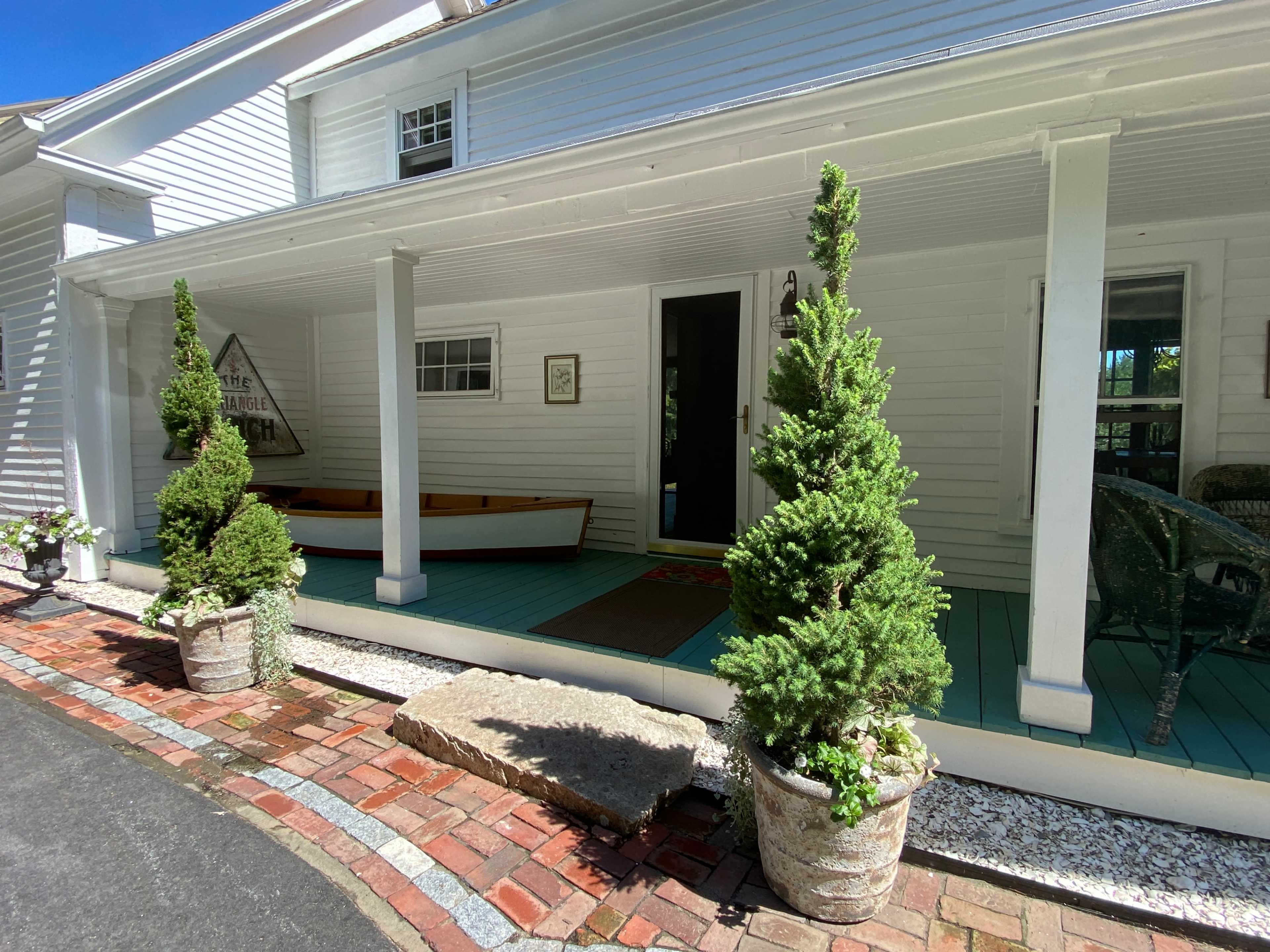 The image shows a white house with a porch, flanked by two potted evergreen trees and a canoe resting on the porch.