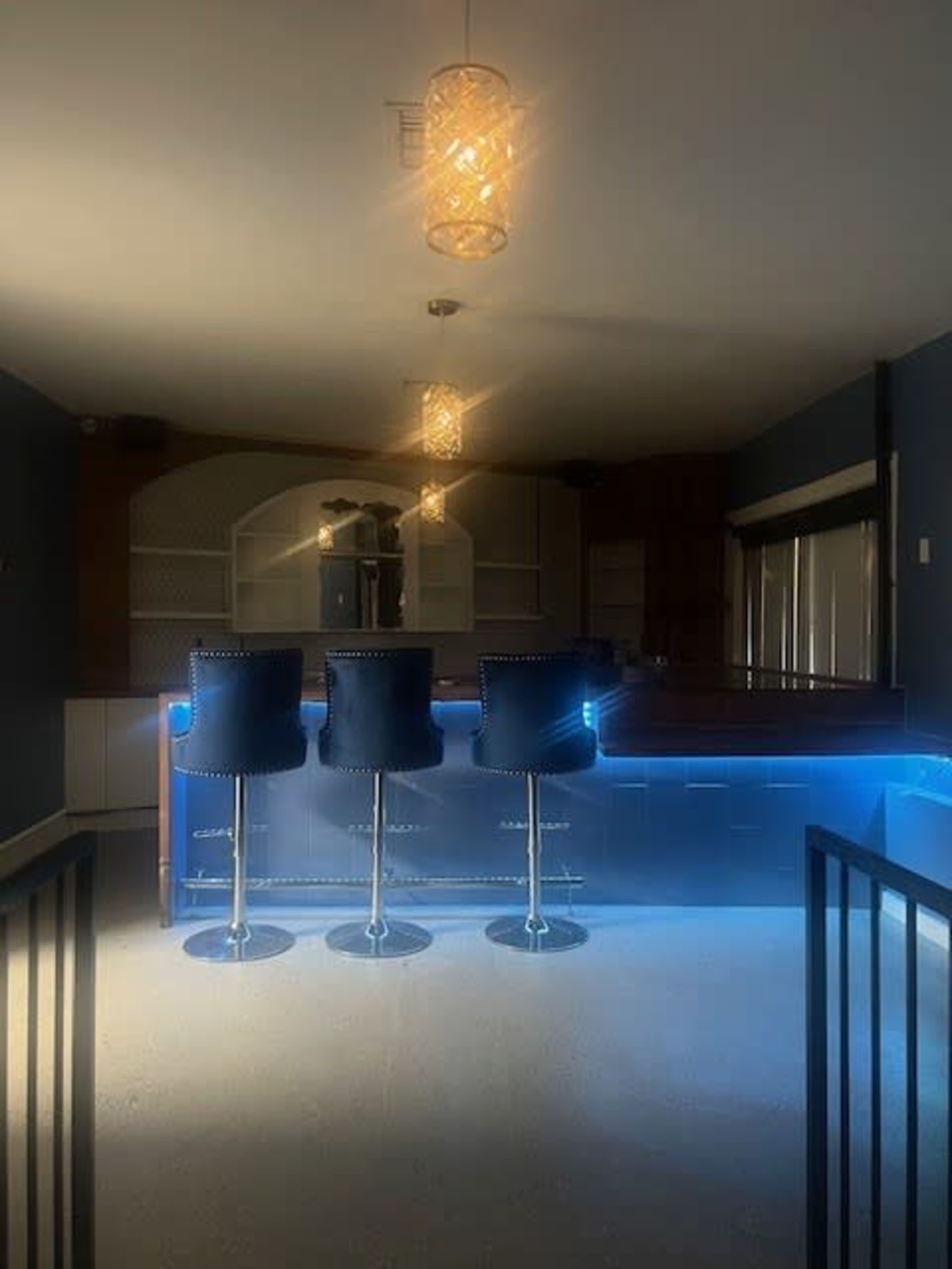 The image shows a modern bar area with three blue stools and illuminated countertops, complemented by hanging light fixtures.