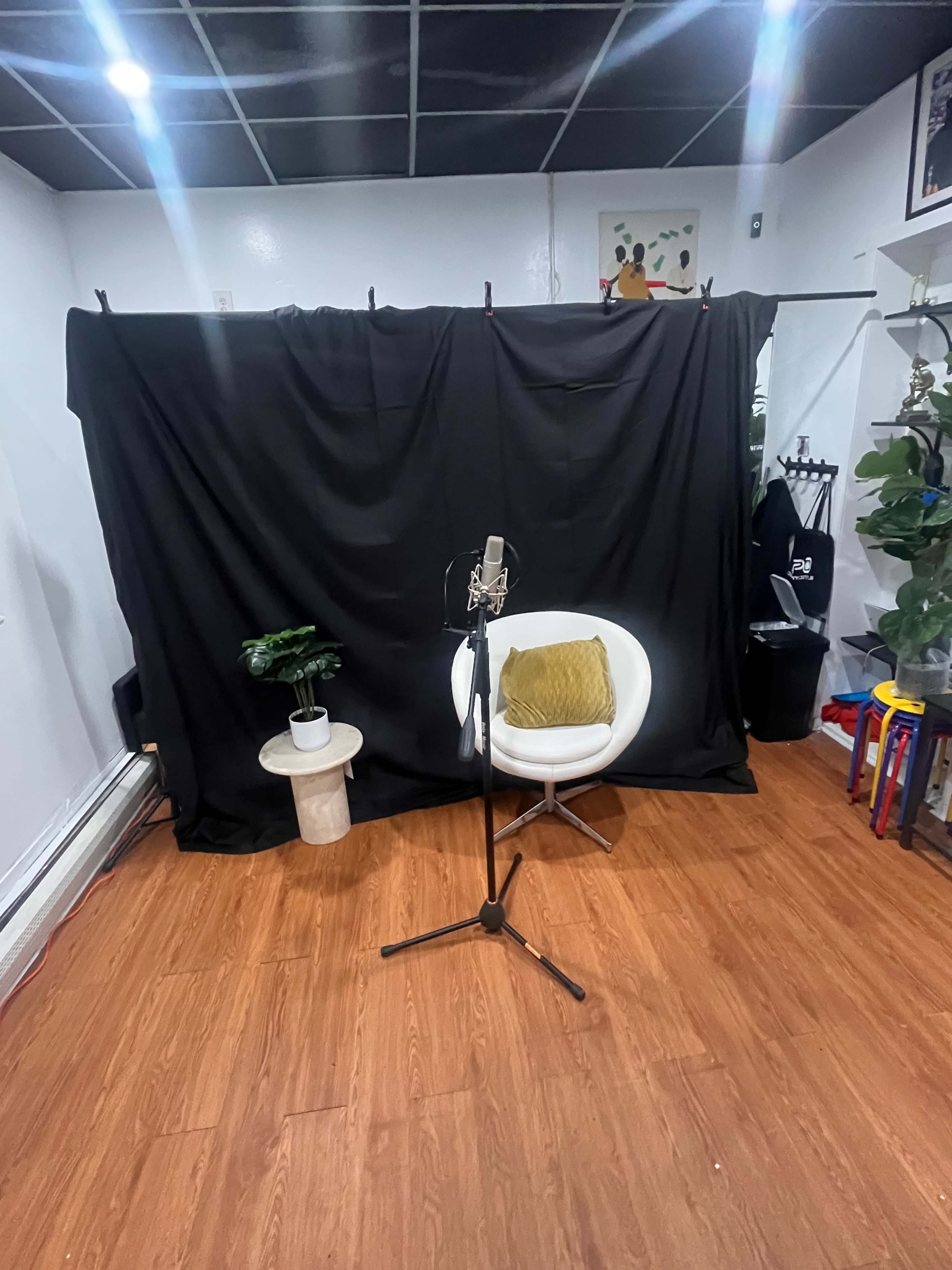 The image shows a small room with a microphone on a stand in front of a white chair, a small table with a plant, and a black curtain draped in the background.