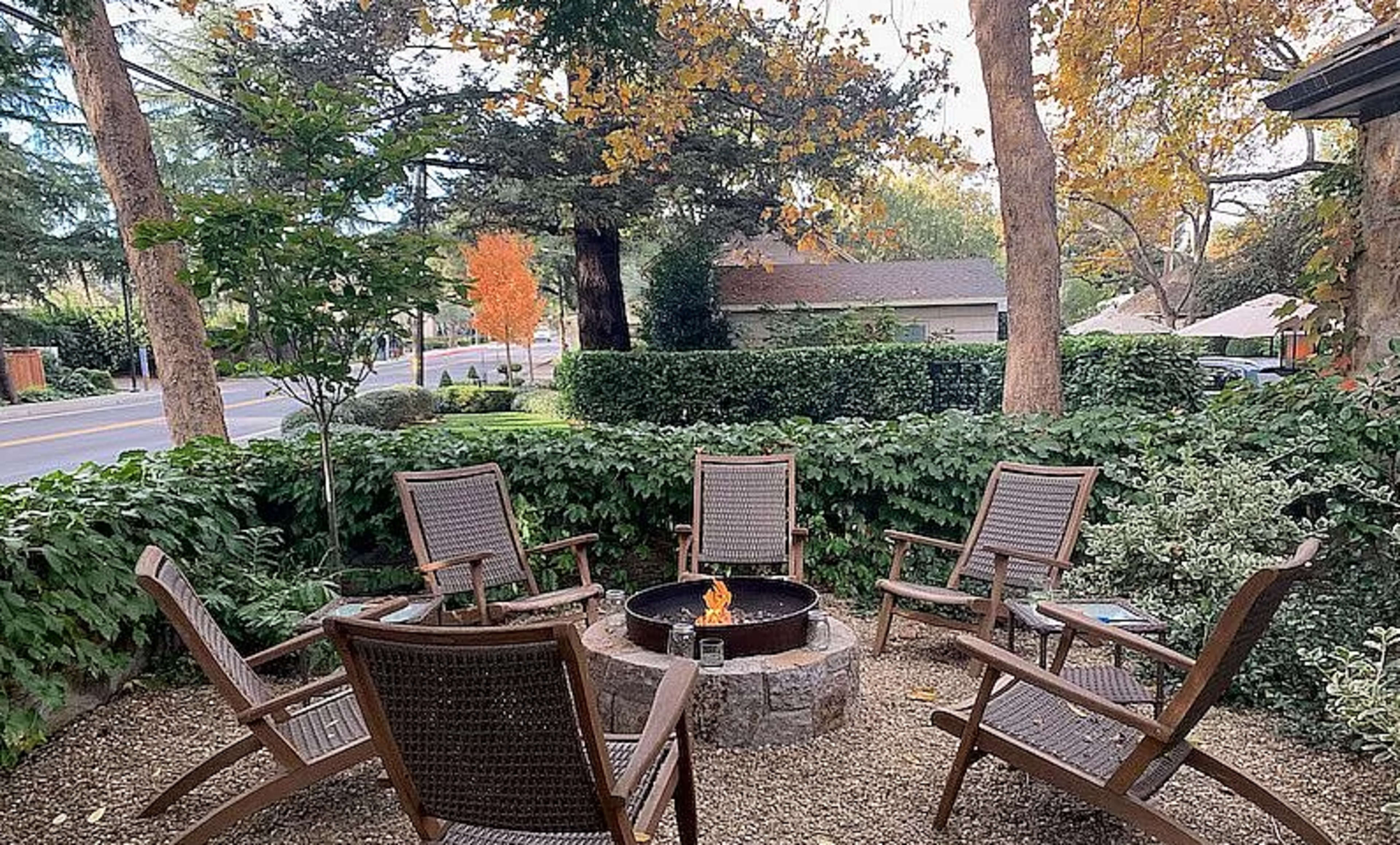 A circular fire pit surrounded by five wooden chairs is set in a landscape of fall-colored trees and lush greenery.