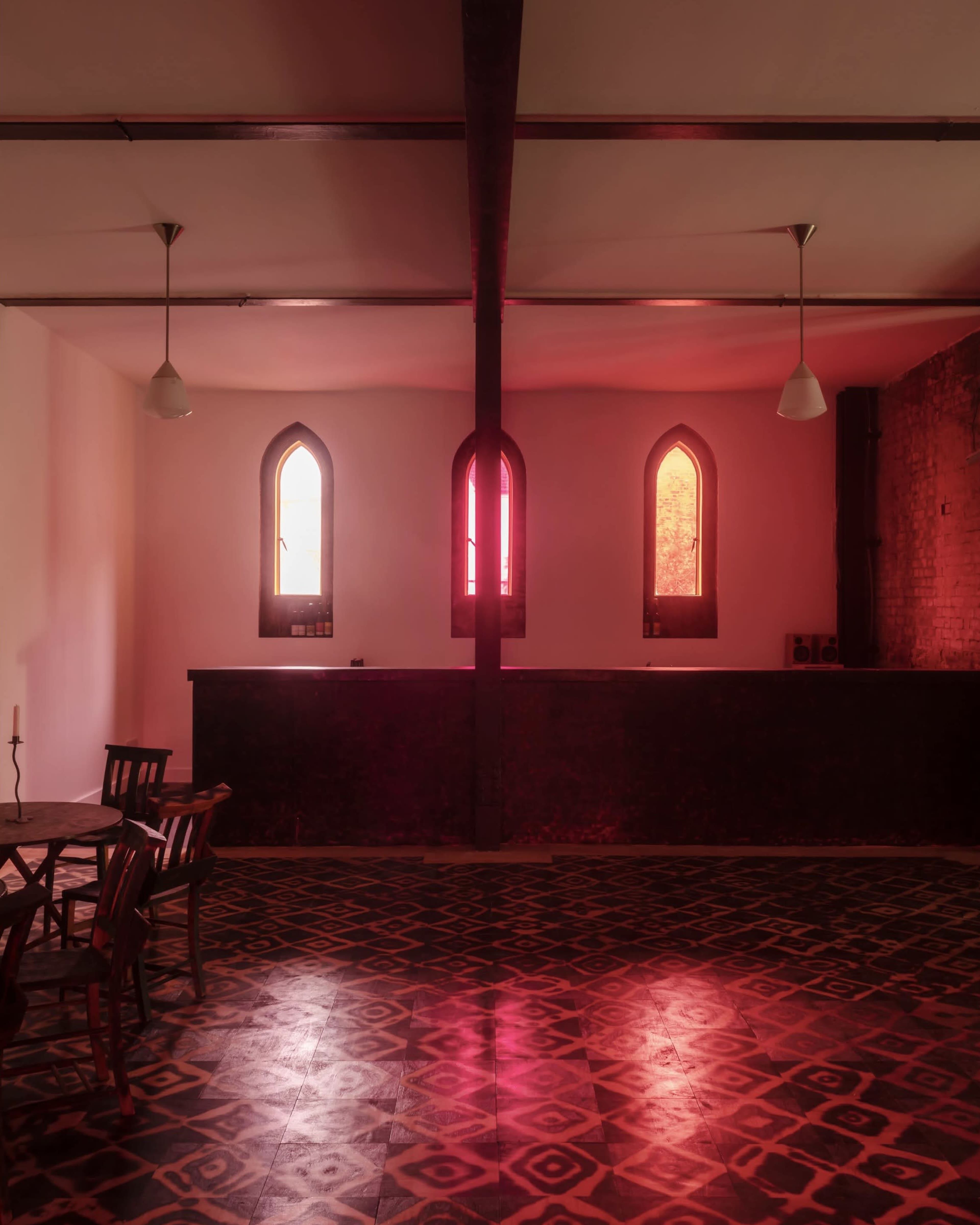 The room features a wooden floor with geometric patterns, a table and chairs, and three arched windows casting red and orange light.