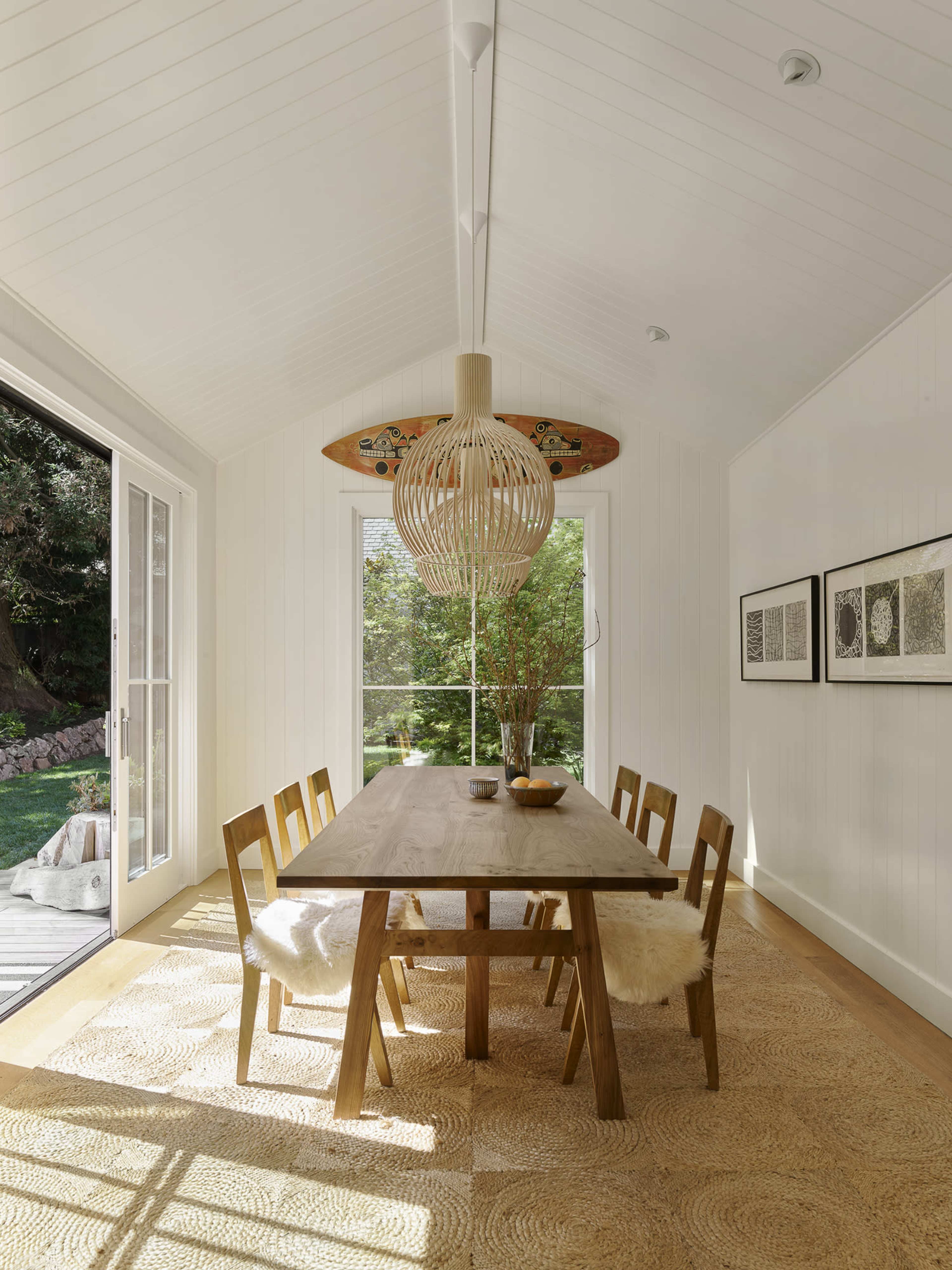 A dining area features a long wooden table surrounded by six chairs, with a large pendant light overhead and windows that let in natural light.