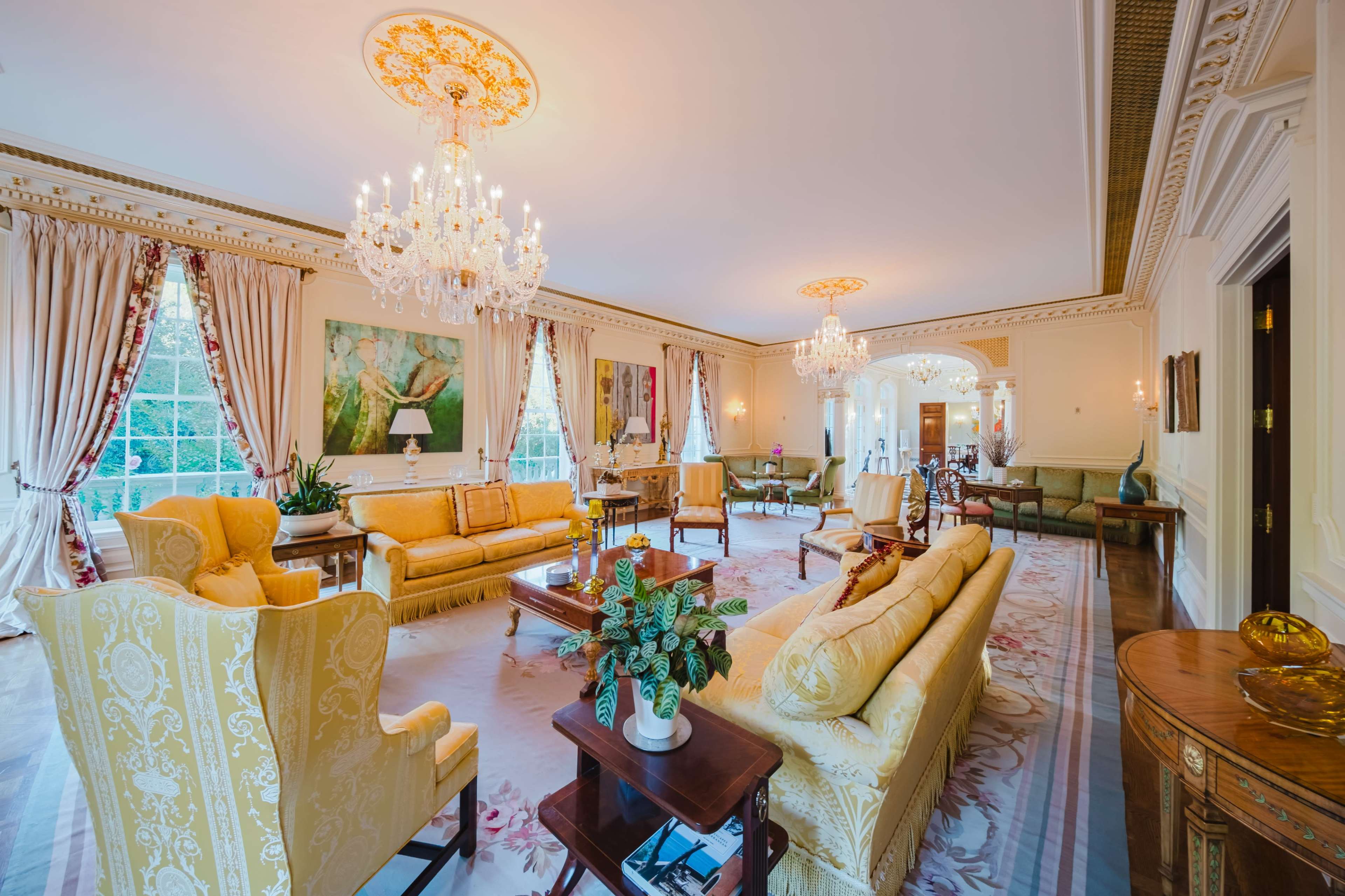 The image shows a spacious, elegantly decorated living room featuring yellow upholstered furniture, crystal chandeliers, and large windows with ornate drapes.