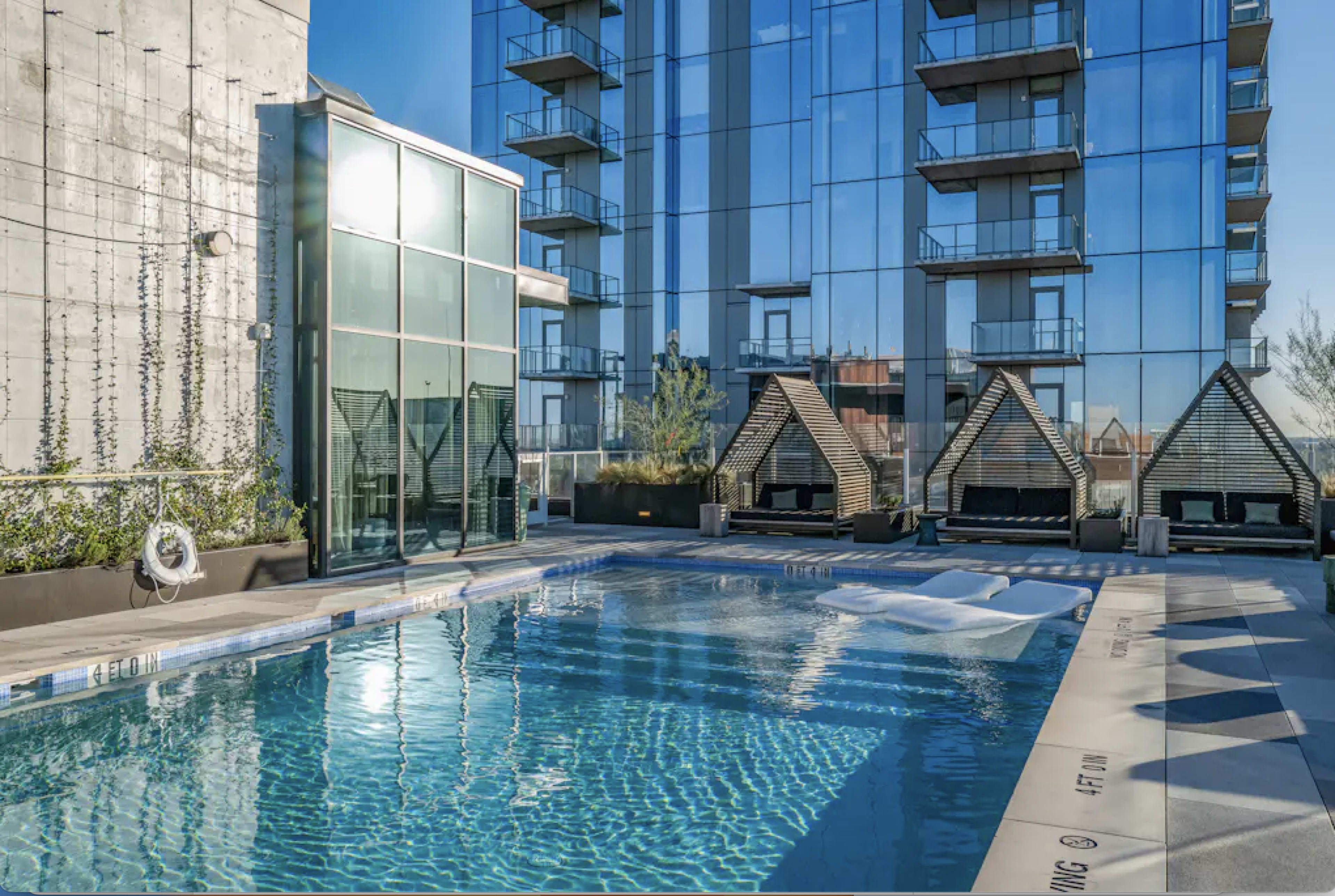 The image shows a modern swimming pool surrounded by lounge areas and a glass building reflecting in the water.