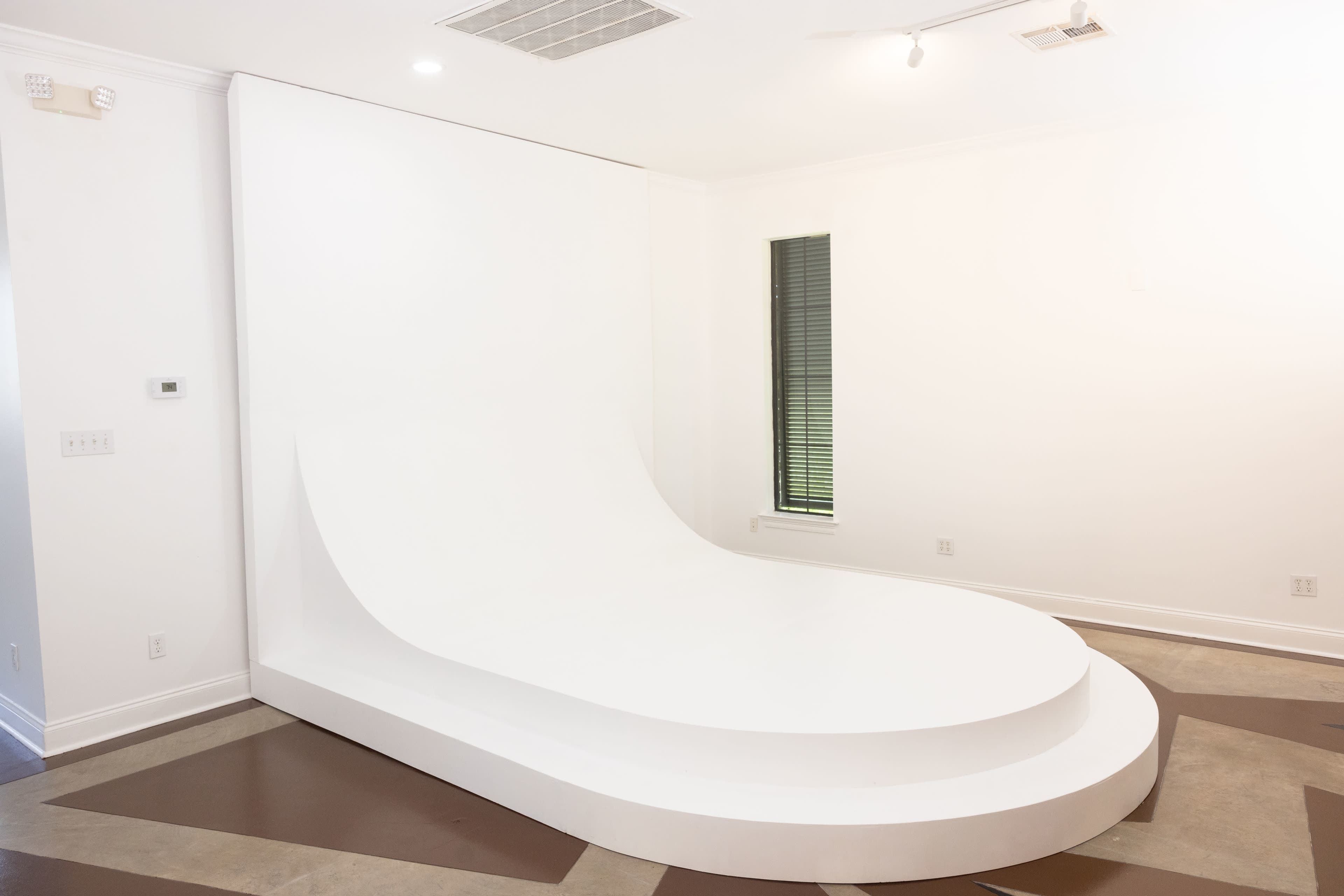 The image shows a minimalist, white curved stage or platform in a well-lit room with large windows.