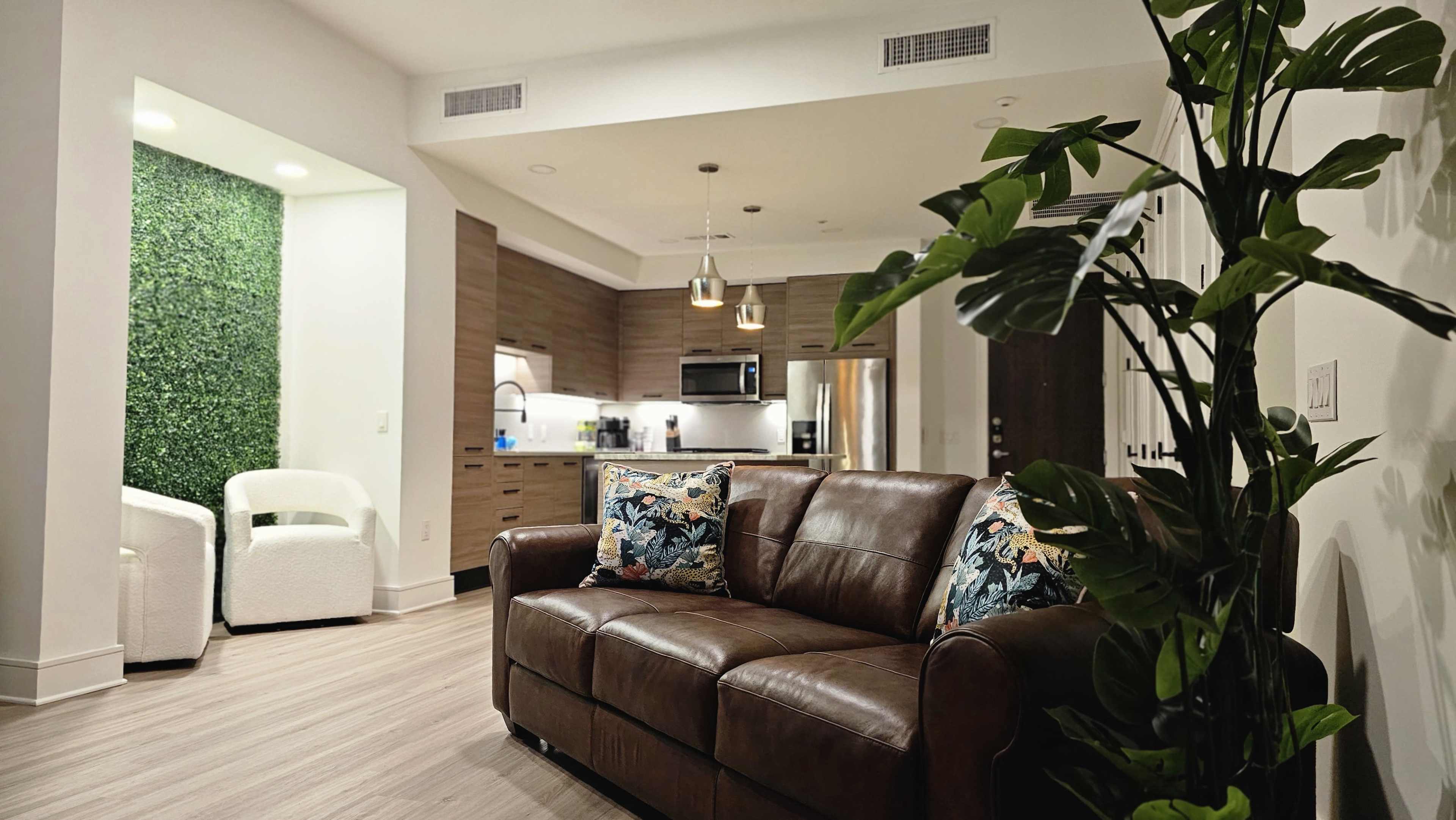 A brown leather couch with decorative pillows is positioned in a modern kitchen and living area that features light wood cabinets and green wall decor.