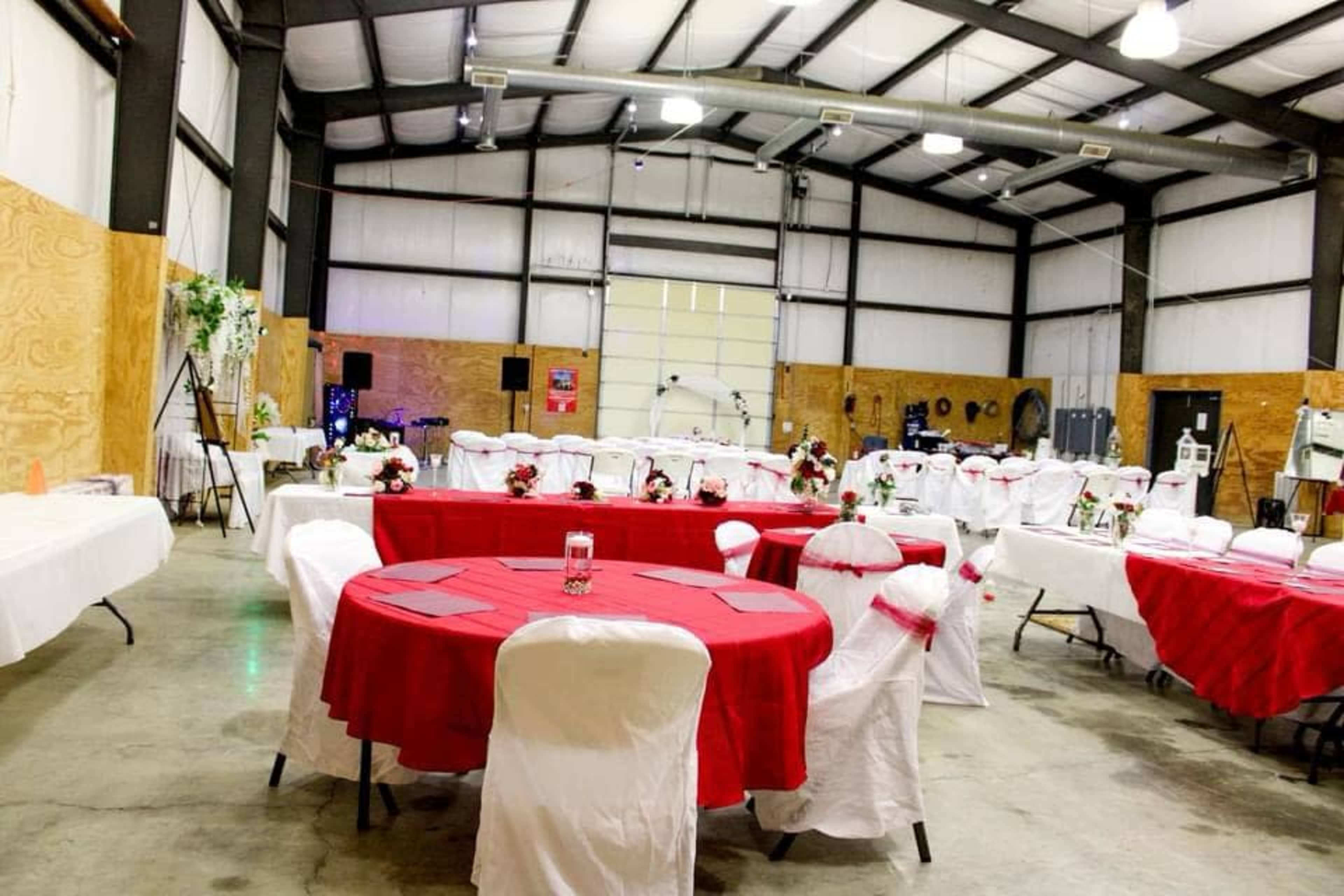 The image shows a large indoor venue set up for an event, featuring tables with red tablecloths and chairs covered in white fabric.