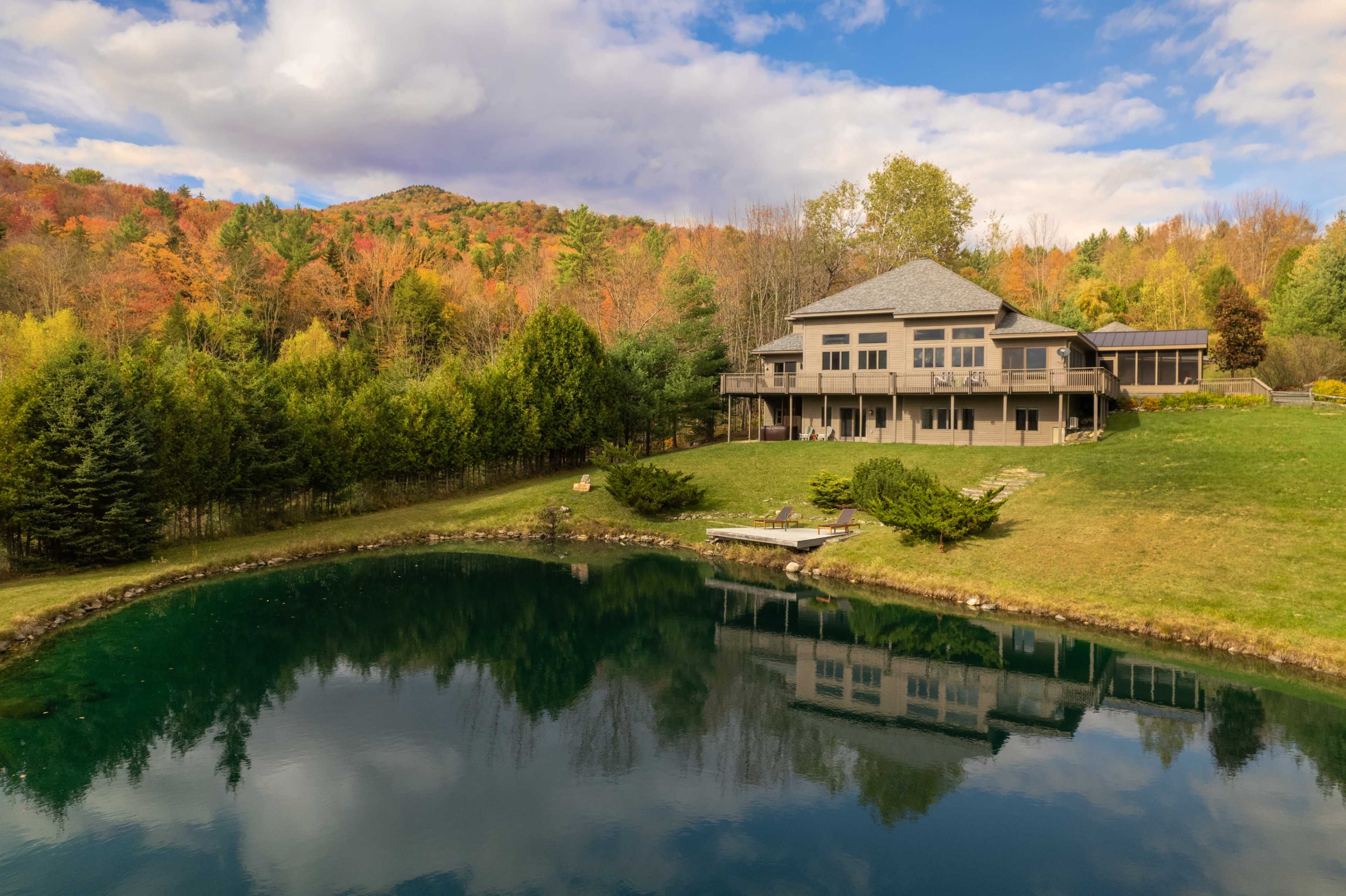 The image shows a large house with a deck overlooking a pond, surrounded by trees with autumn foliage and a mountainous background.