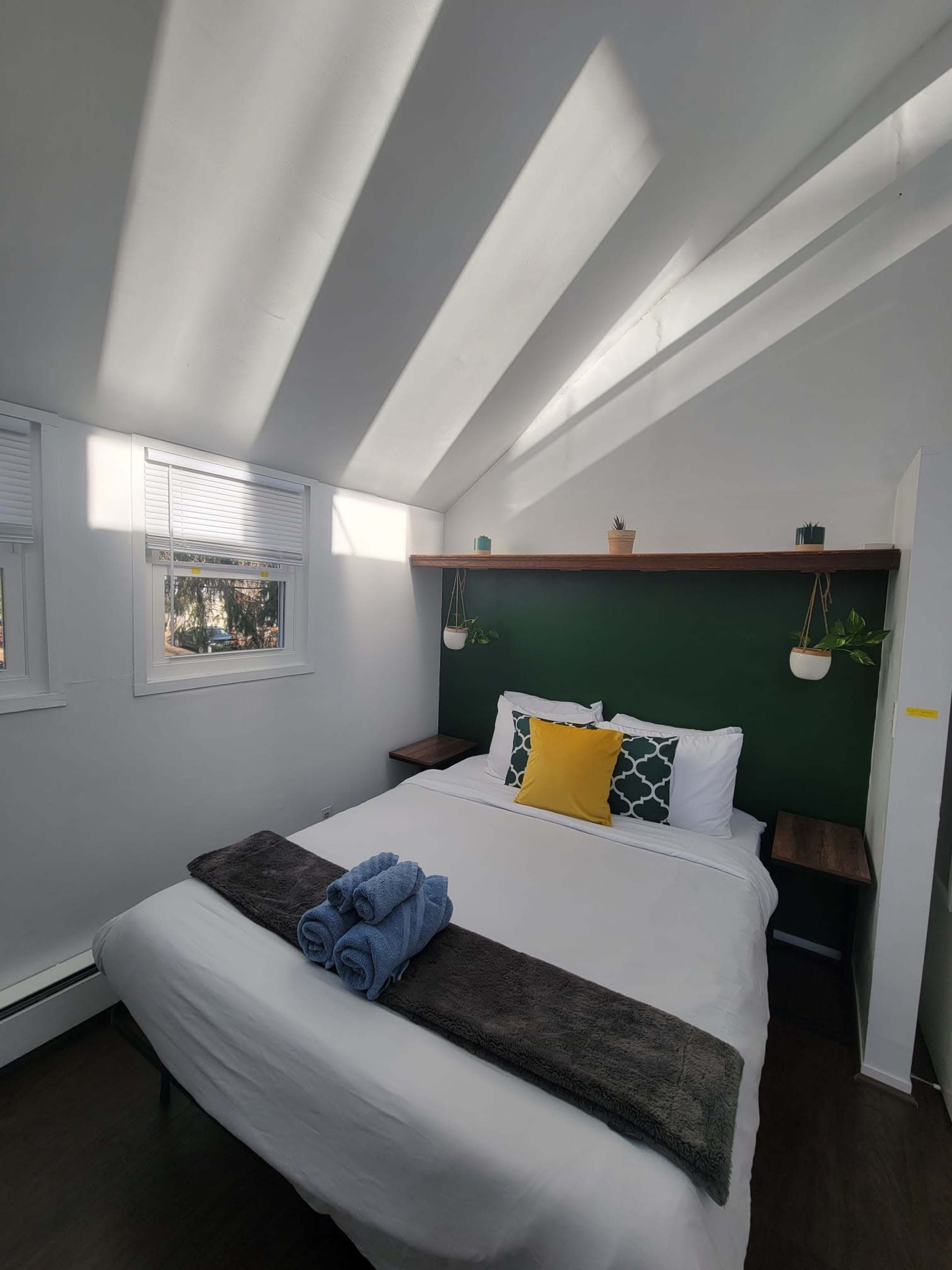 A white bedroom features a bed with a yellow cushion, a dark throw, and rolled towels, with green walls and natural light streaming through the windows.