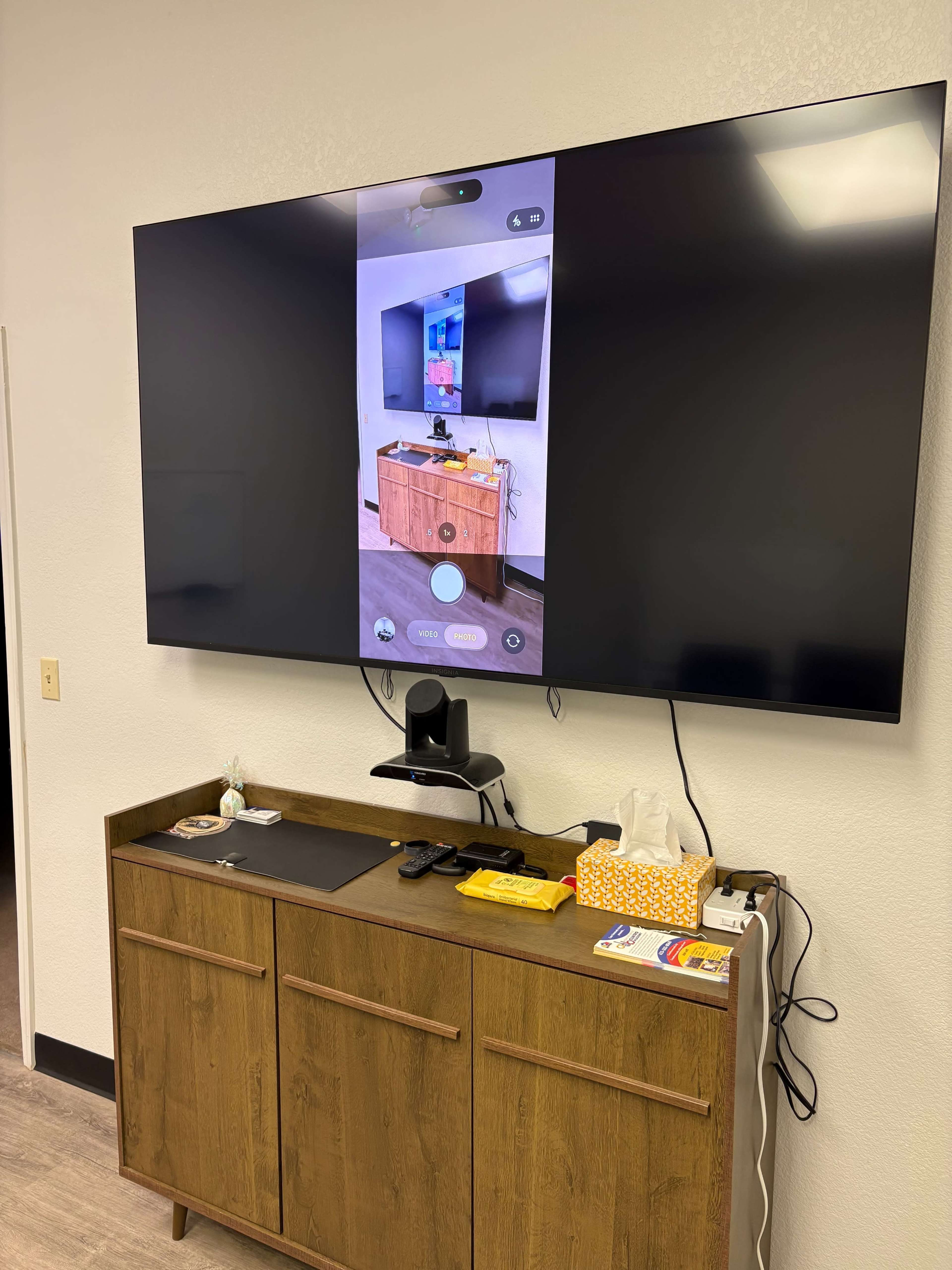 The image shows a large television mounted on a wall displaying a digital camera view of a room, with a wooden cabinet underneath containing various items.