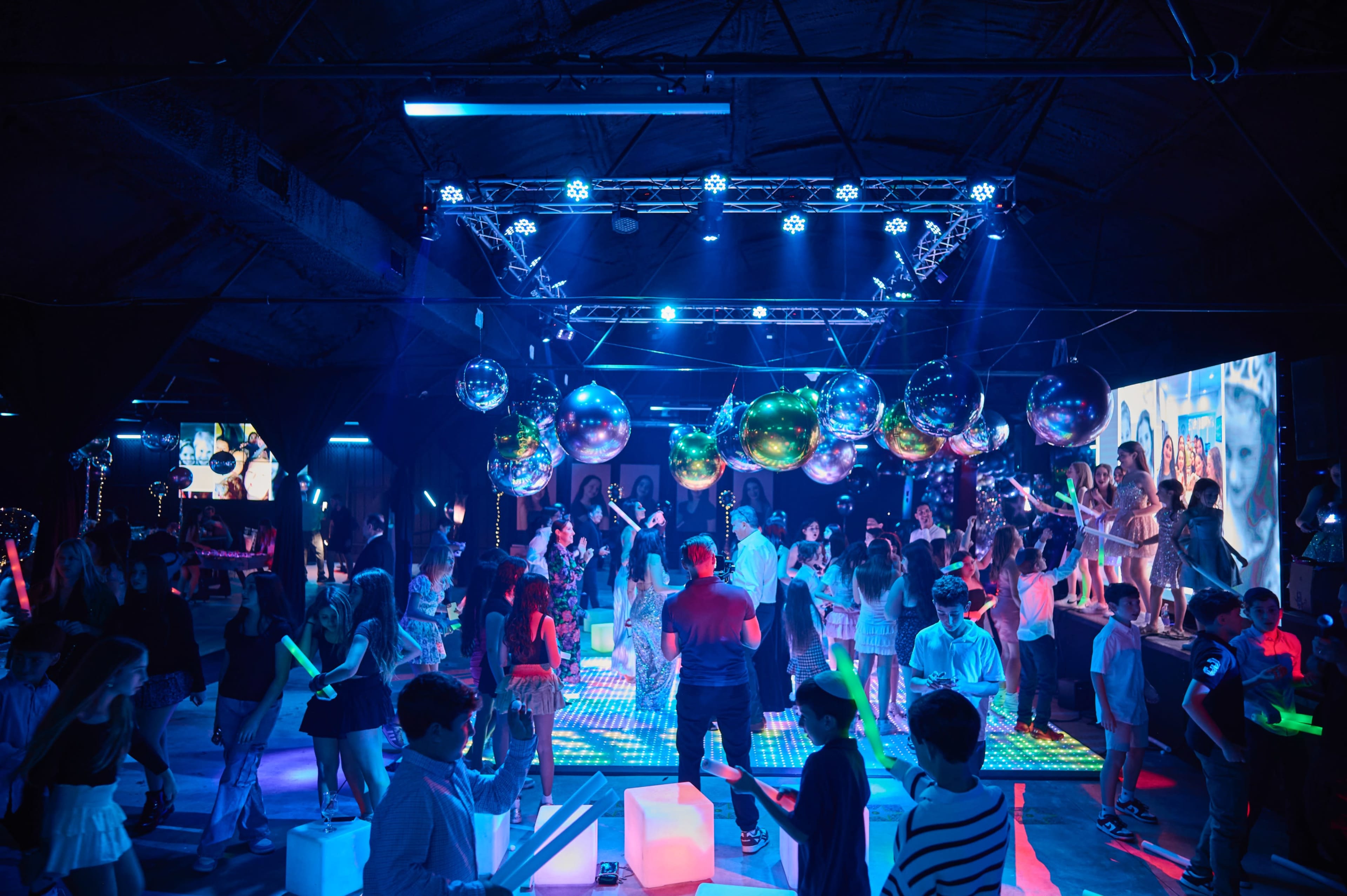 A vibrant dance party features numerous people dancing under colorful lights and disco balls in a dimly lit venue.