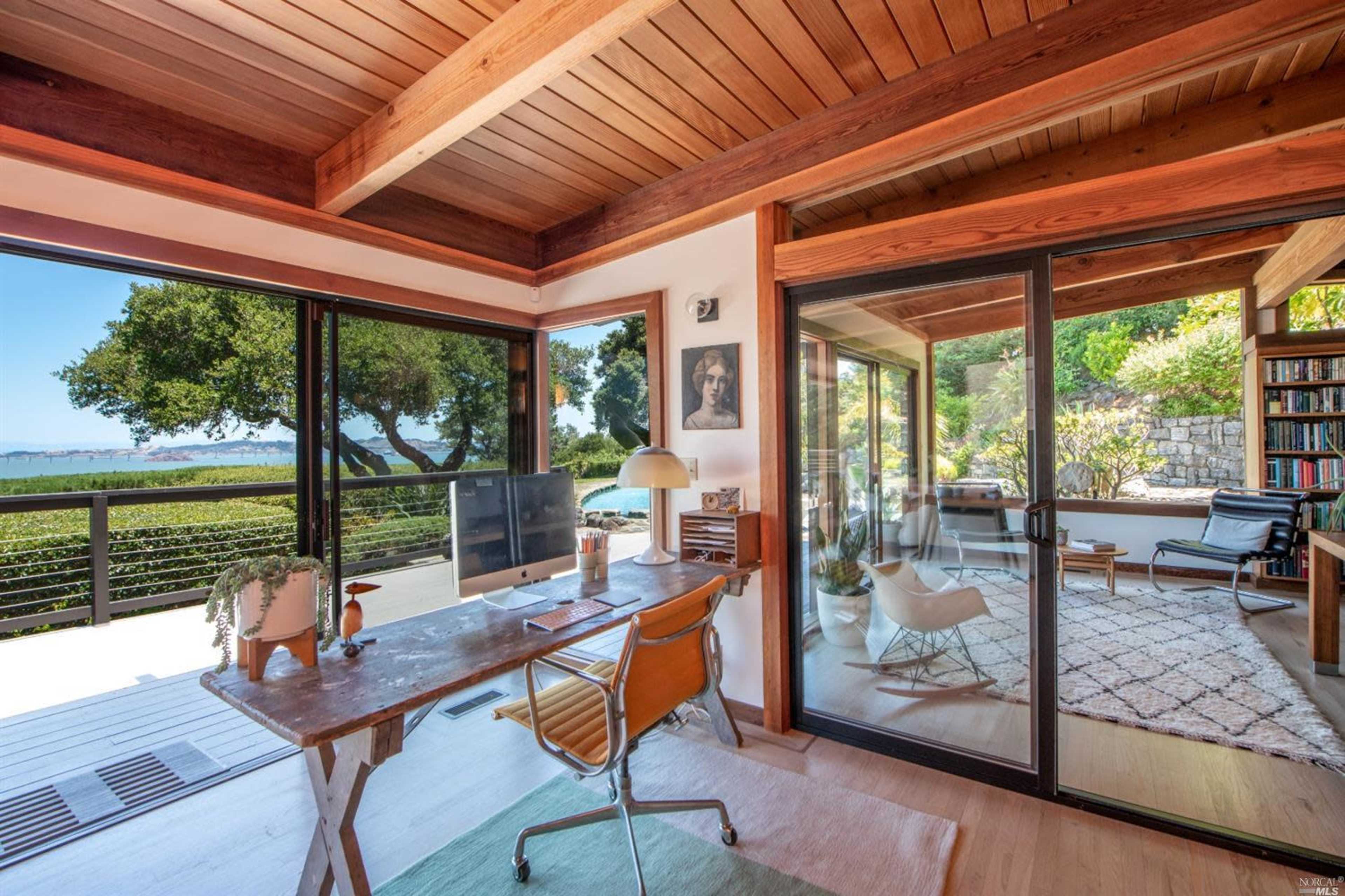 A modern home office features a wooden desk and chair, surrounded by large glass windows offering a view of lush greenery and a distant landscape.