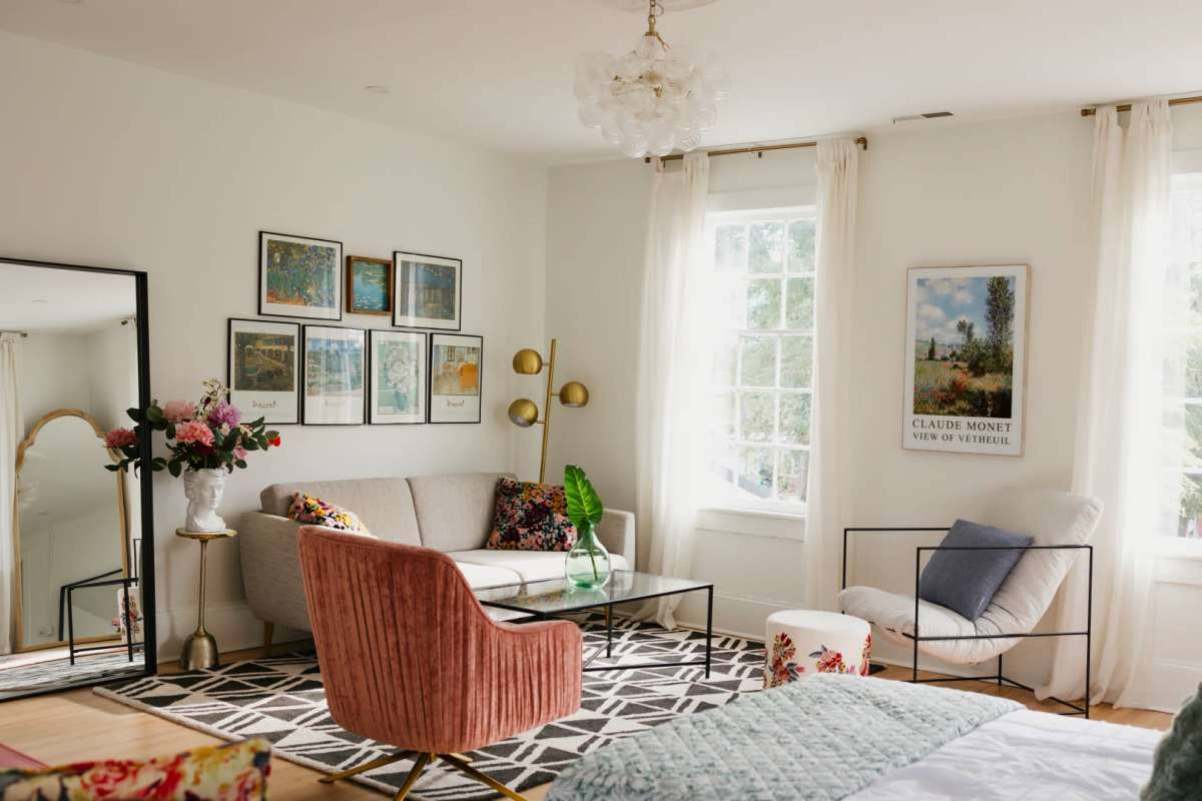 The image shows a cozy living room featuring a sofa, armchair, decorative art on the walls, and natural light streaming through large windows.