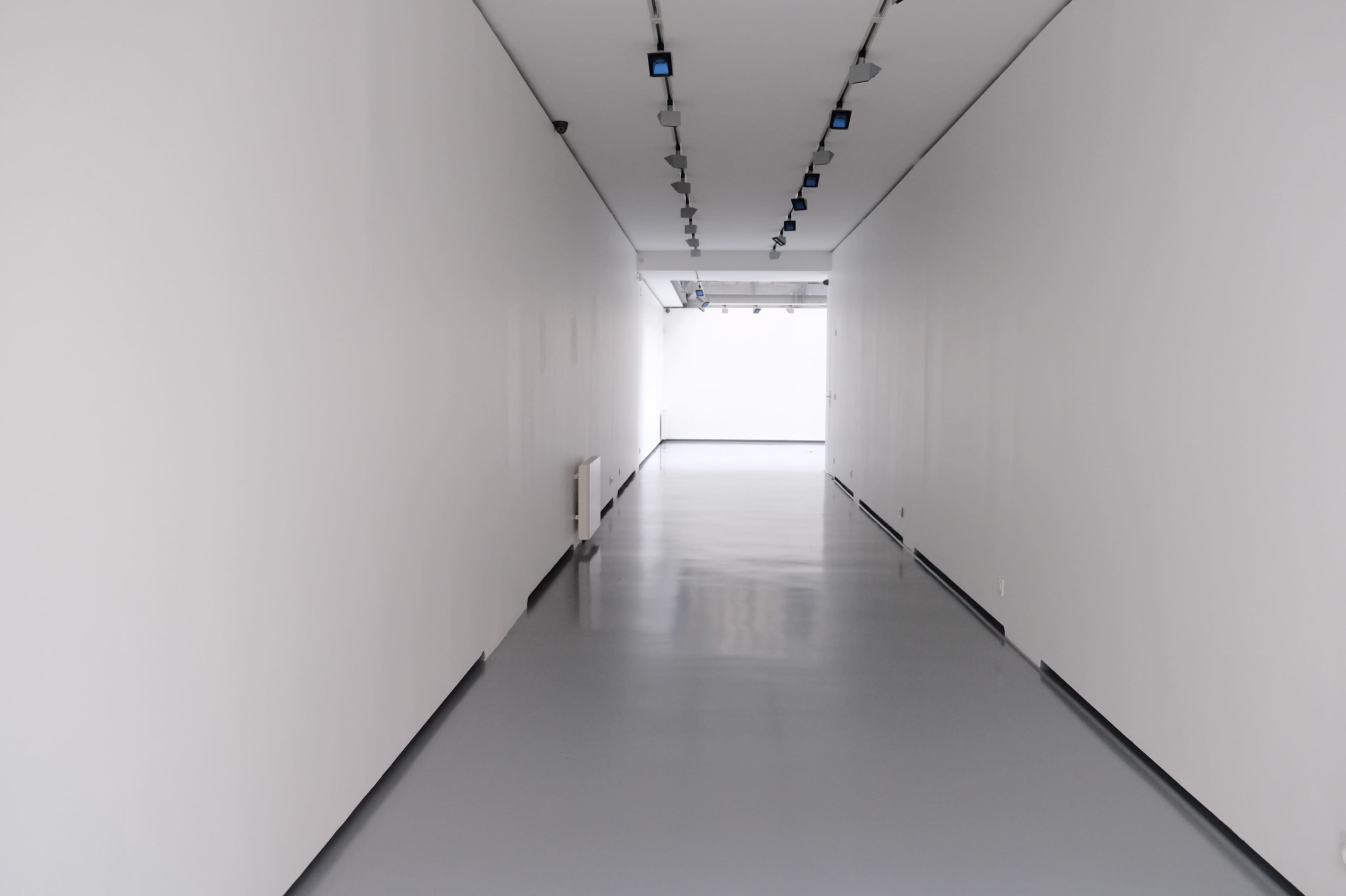 The image shows a long, narrow hallway with white walls and a smooth gray floor, illuminated by ceiling lights.