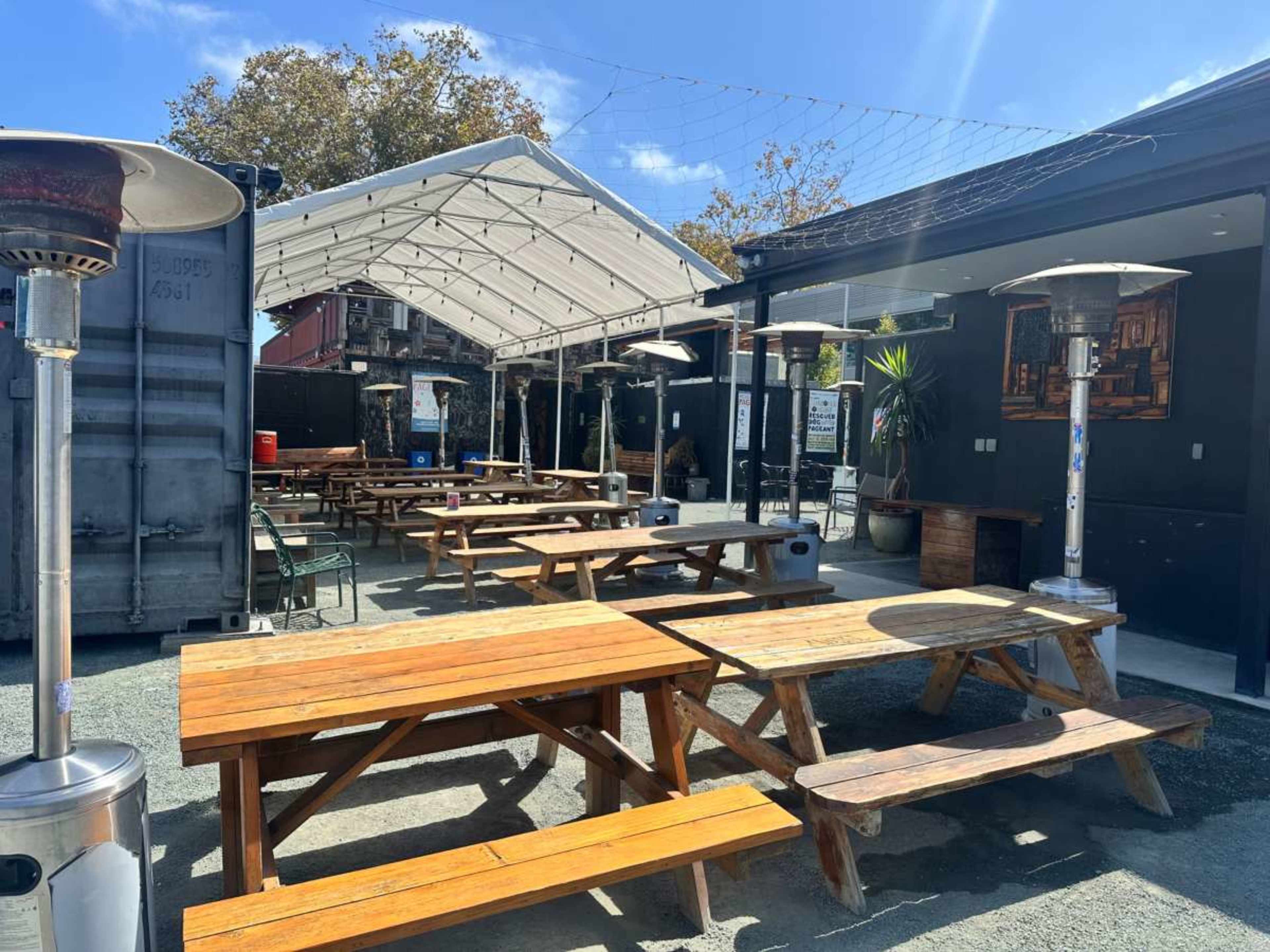The image shows an outdoor dining area with several wooden picnic tables, patio heaters, and a canopy covering the space, set in a gravel surface.