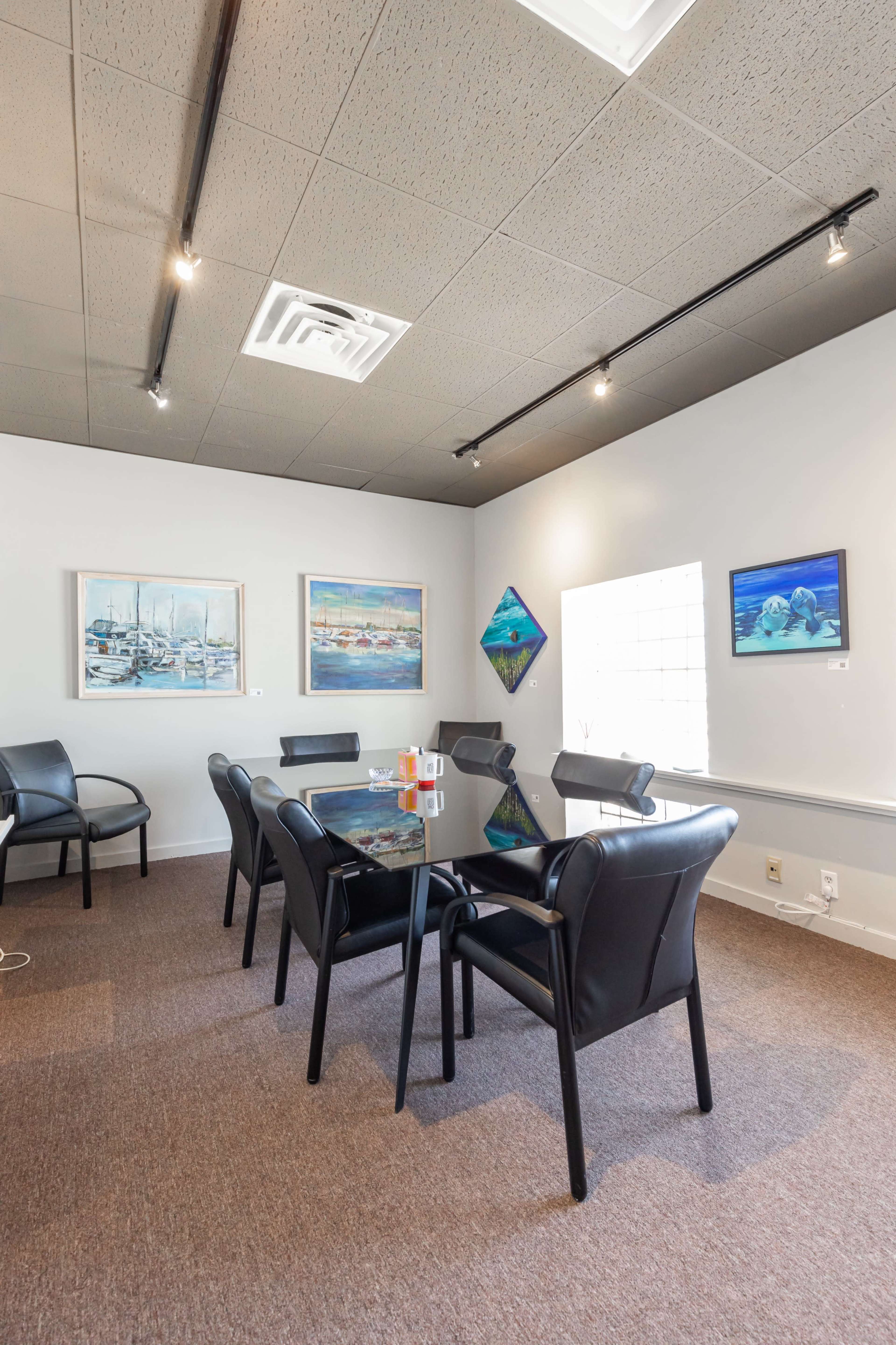 A conference room with a glass table surrounded by black chairs, adorned with paintings on the walls and natural light coming through a window.