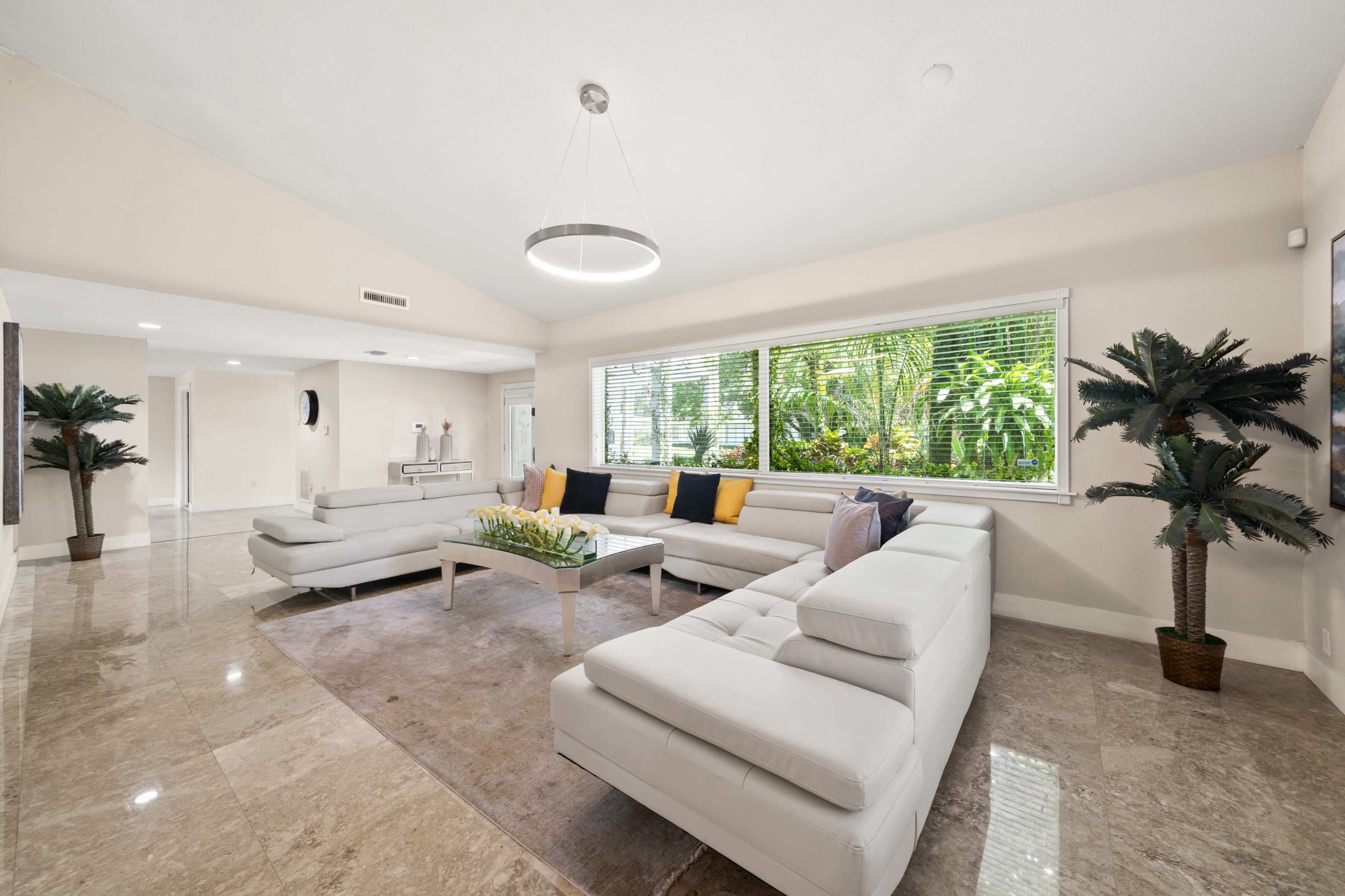 A modern living room features a sectional sofa, a coffee table with decorative items, and large windows overlooking greenery.